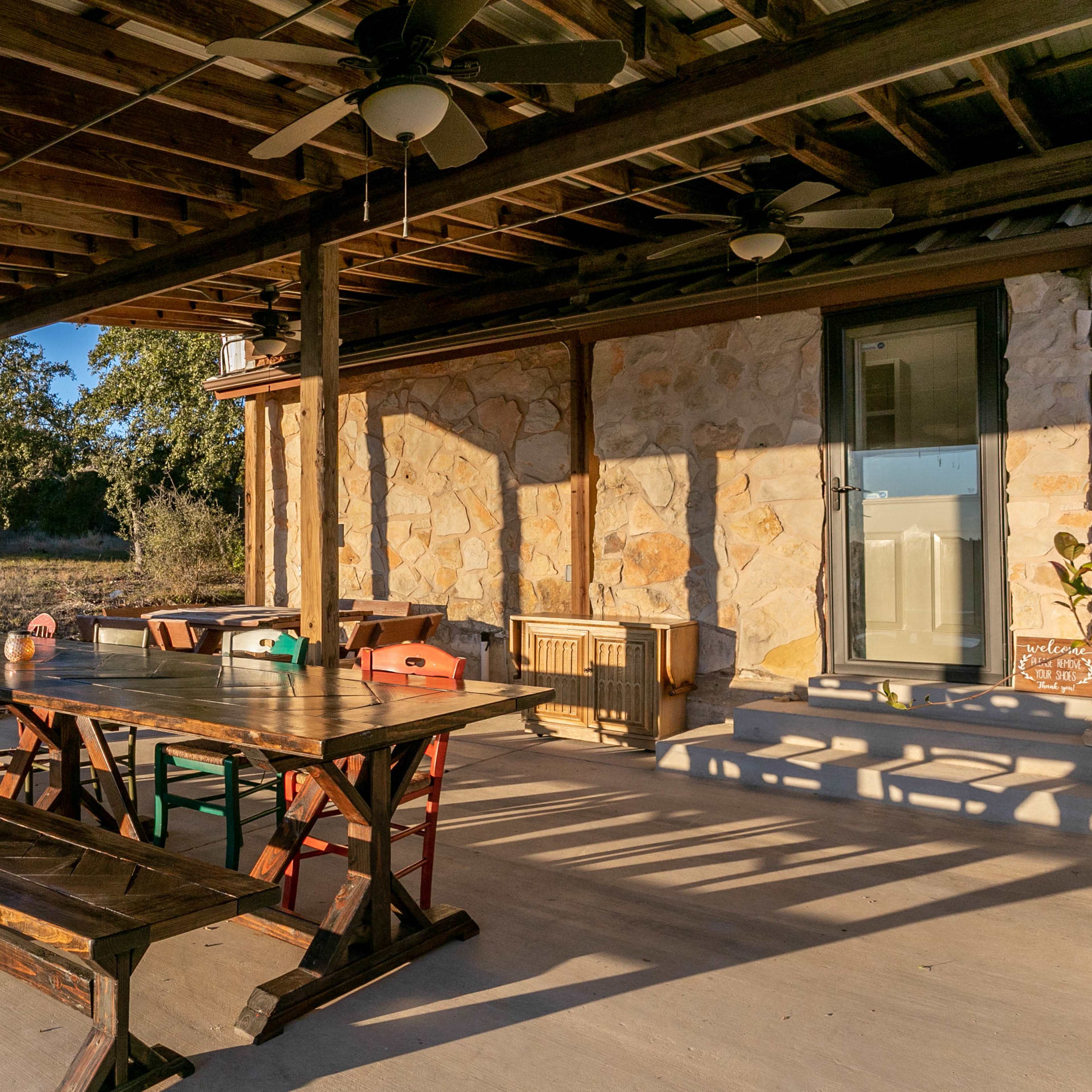 A covered patio features a large wooden table with benches, ceiling fans, and a stone wall, set in a rural landscape.