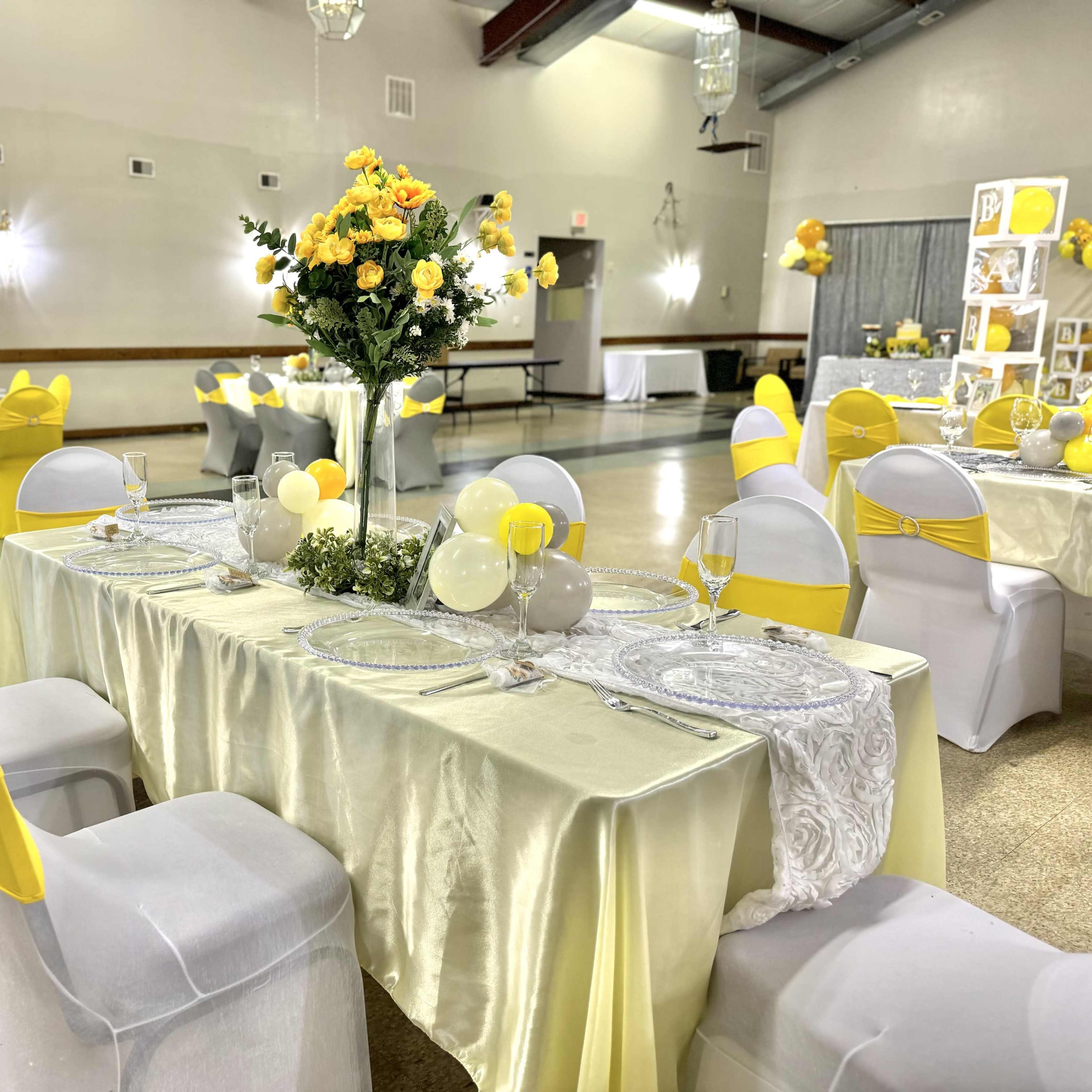 A banquet hall is set up with tables dressed in light yellow tablecloths, adorned with floral centerpieces and decorated with yellow accents, while chairs are covered in white with yellow sashes.