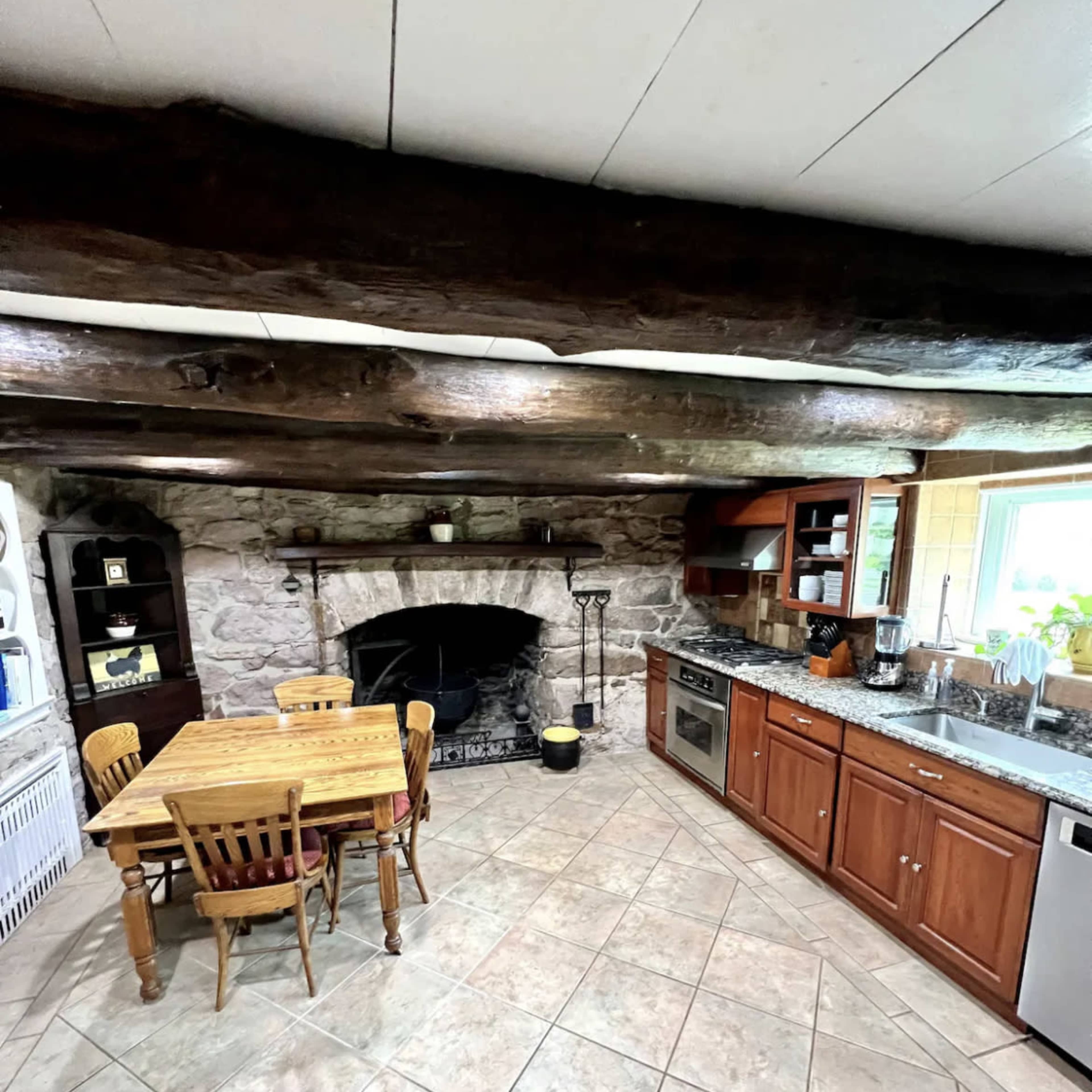 The kitchen features a stone fireplace, wooden beams, a tiled floor, and a dining table surrounded by chairs.