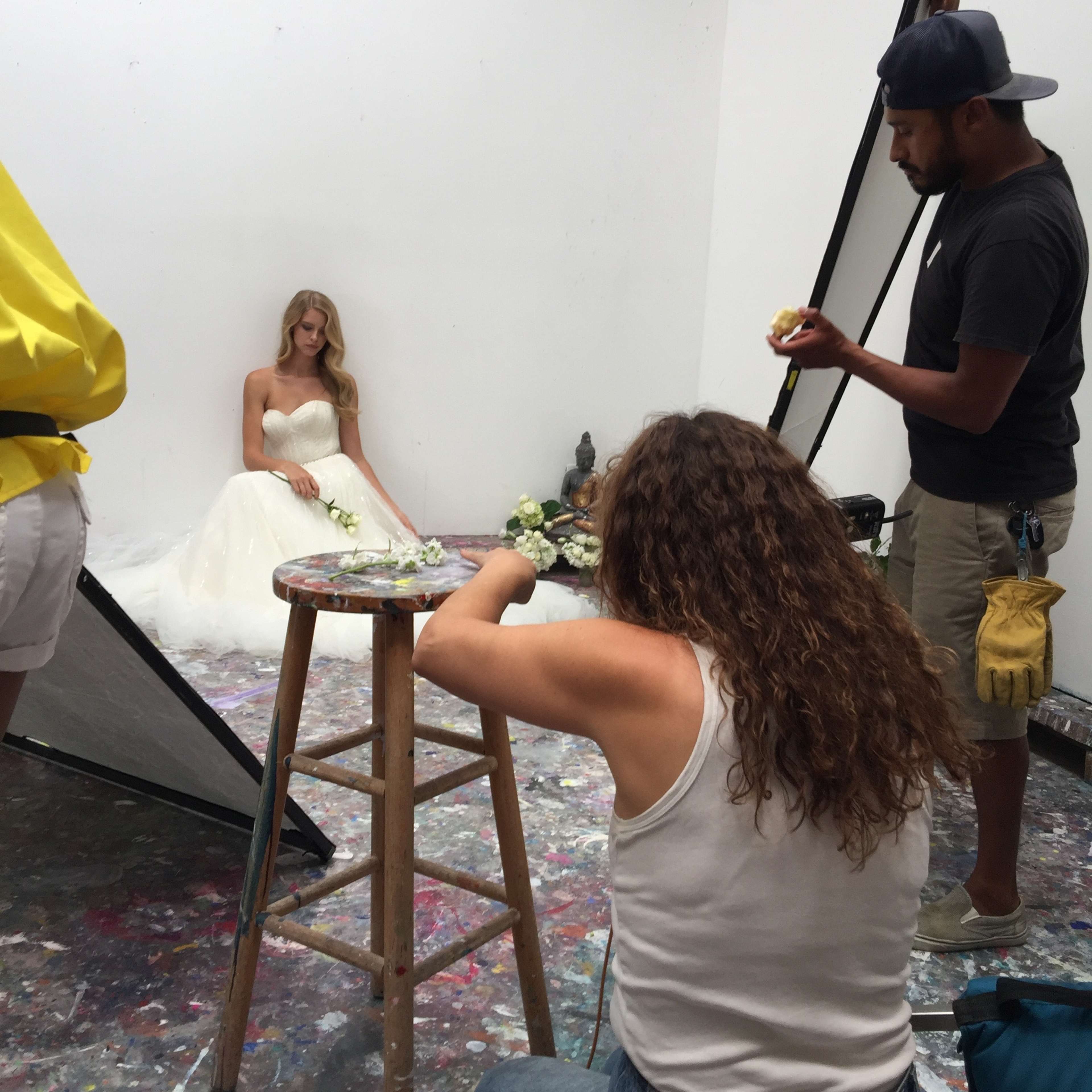 A model in a wedding dress sits against the wall while two people work on a photography setup in a studio filled with colorful splatters on the floor.