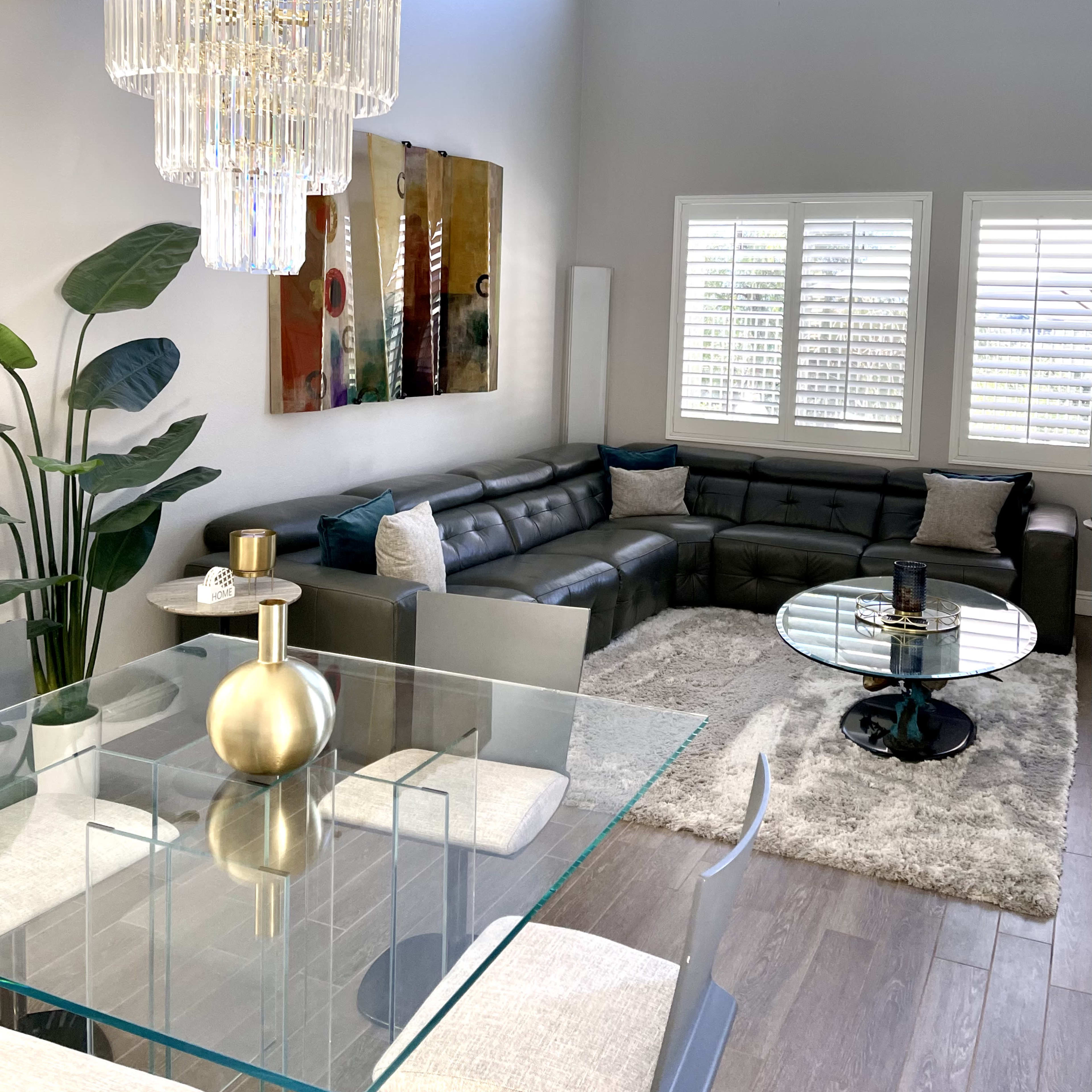 A modern living room and dining area featuring a glass table, a sectional couch, a chandelier, and large windows with plantation shutters.