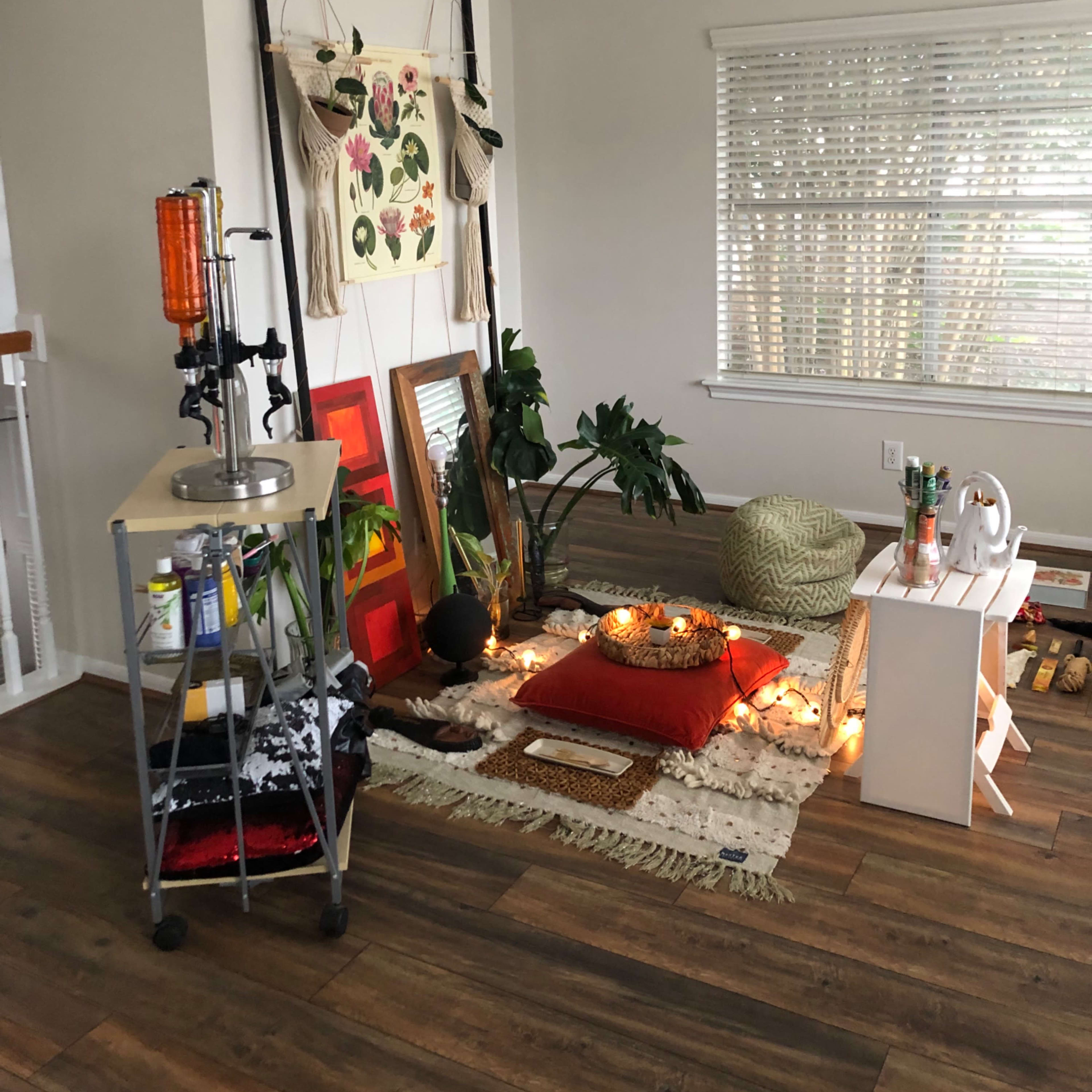 A cozy corner of a room features a relaxed seating area with cushions, plants, and decorative items, illuminated by string lights.