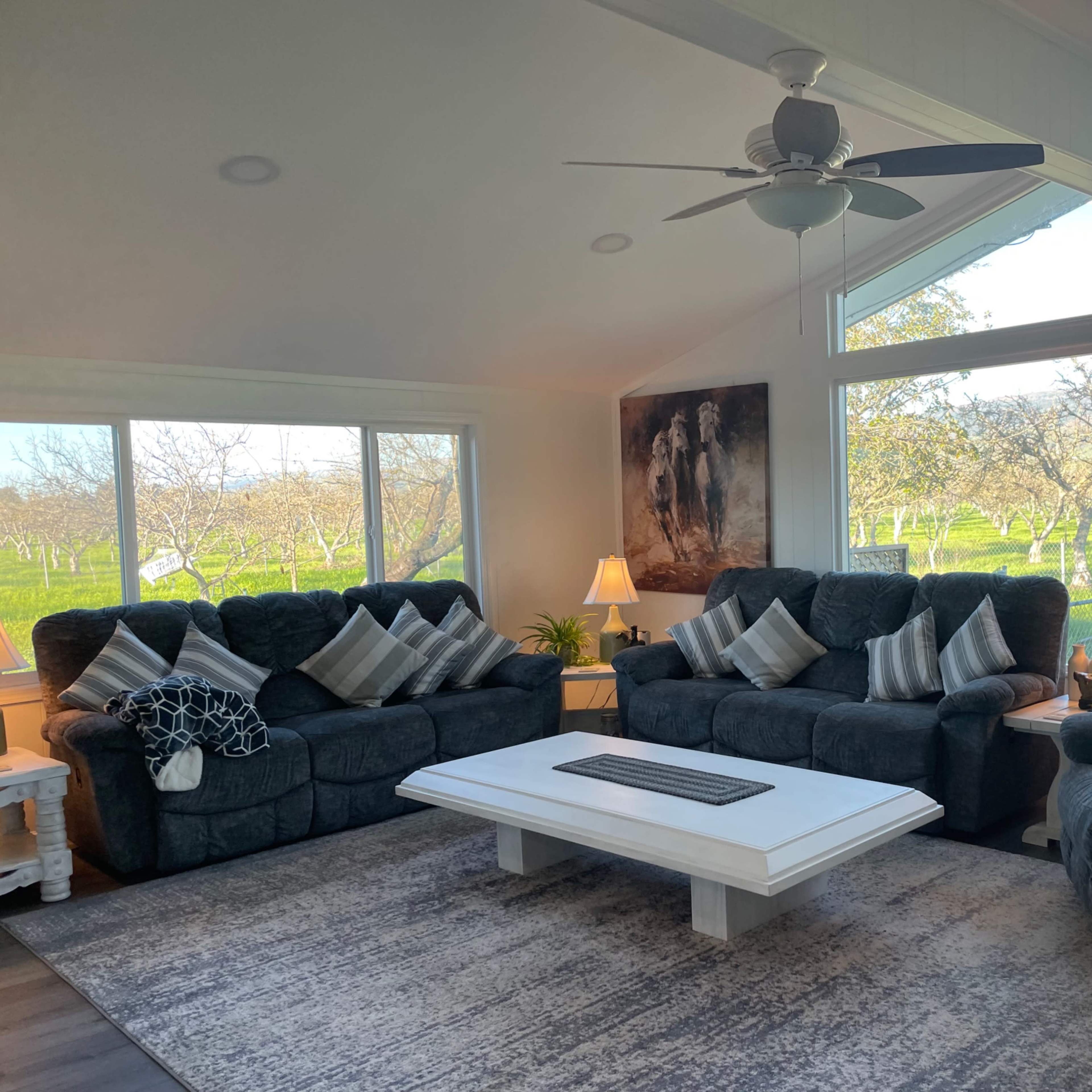 A modern living room features two dark sofas and a coffee table, with large windows overlooking an orchard.
