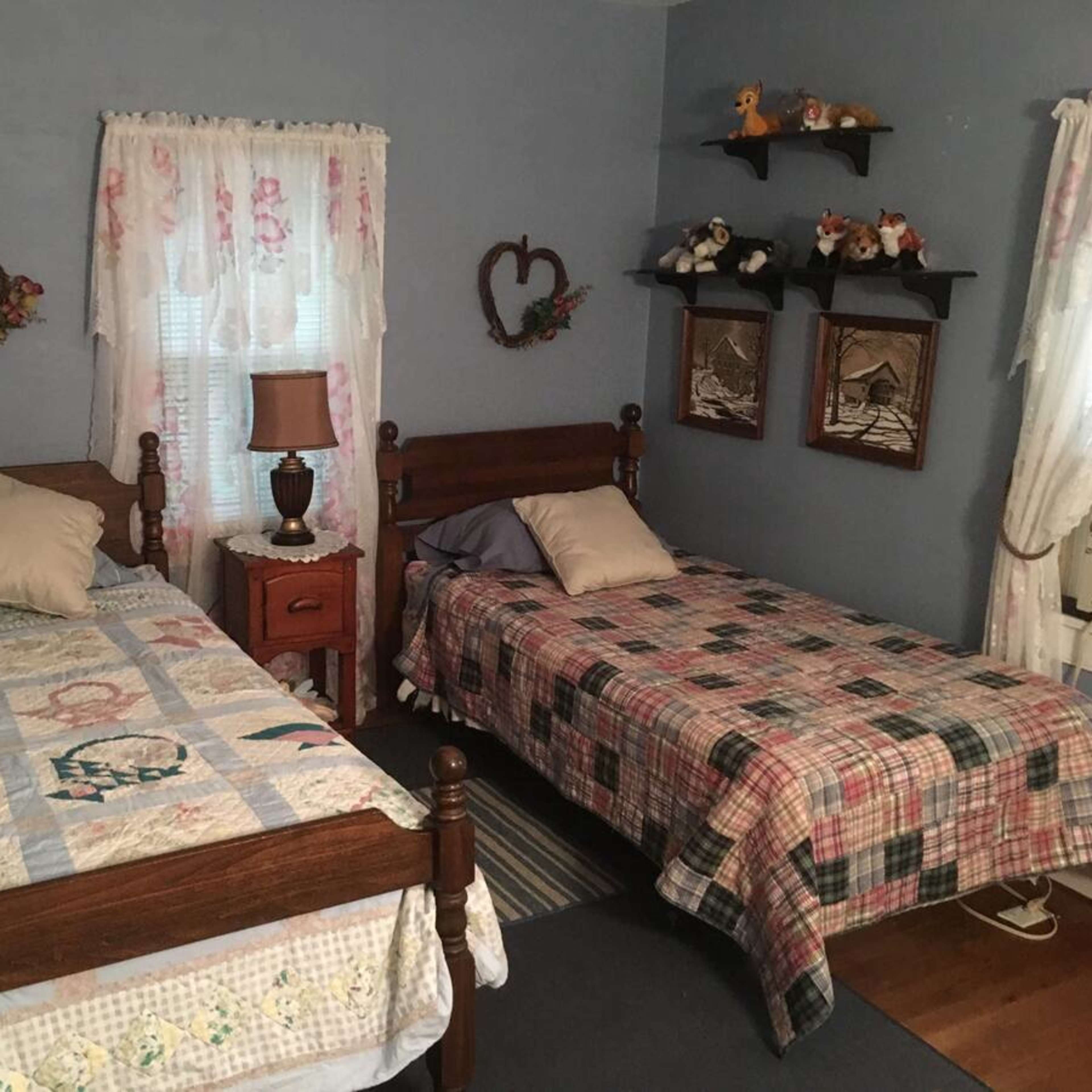 A small guest room featuring two wooden twin beds with colorful quilts, a bedside table with a lamp, and decorative wall accents.