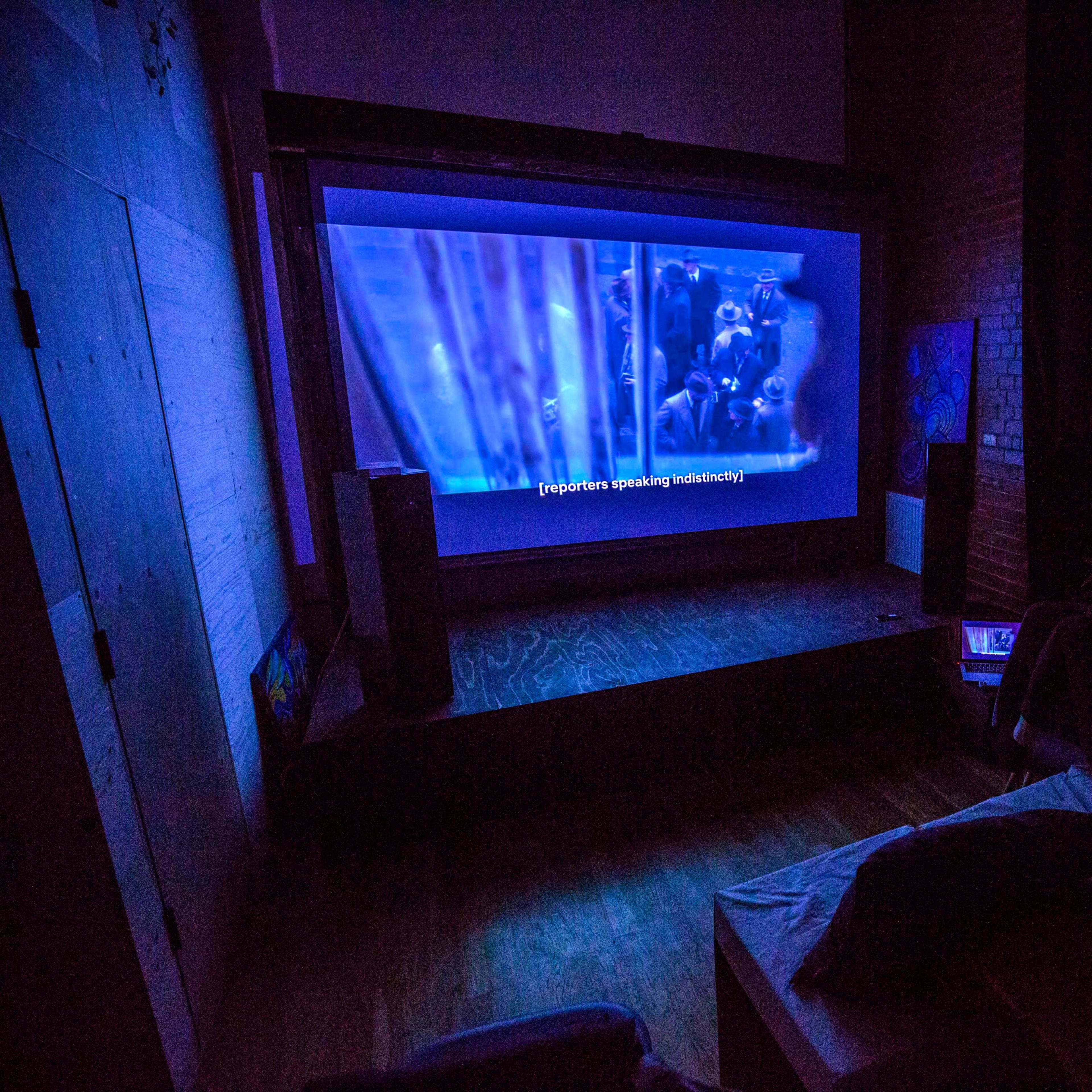 A person is sitting on a couch watching a movie projected on a large screen in a dimly lit room.