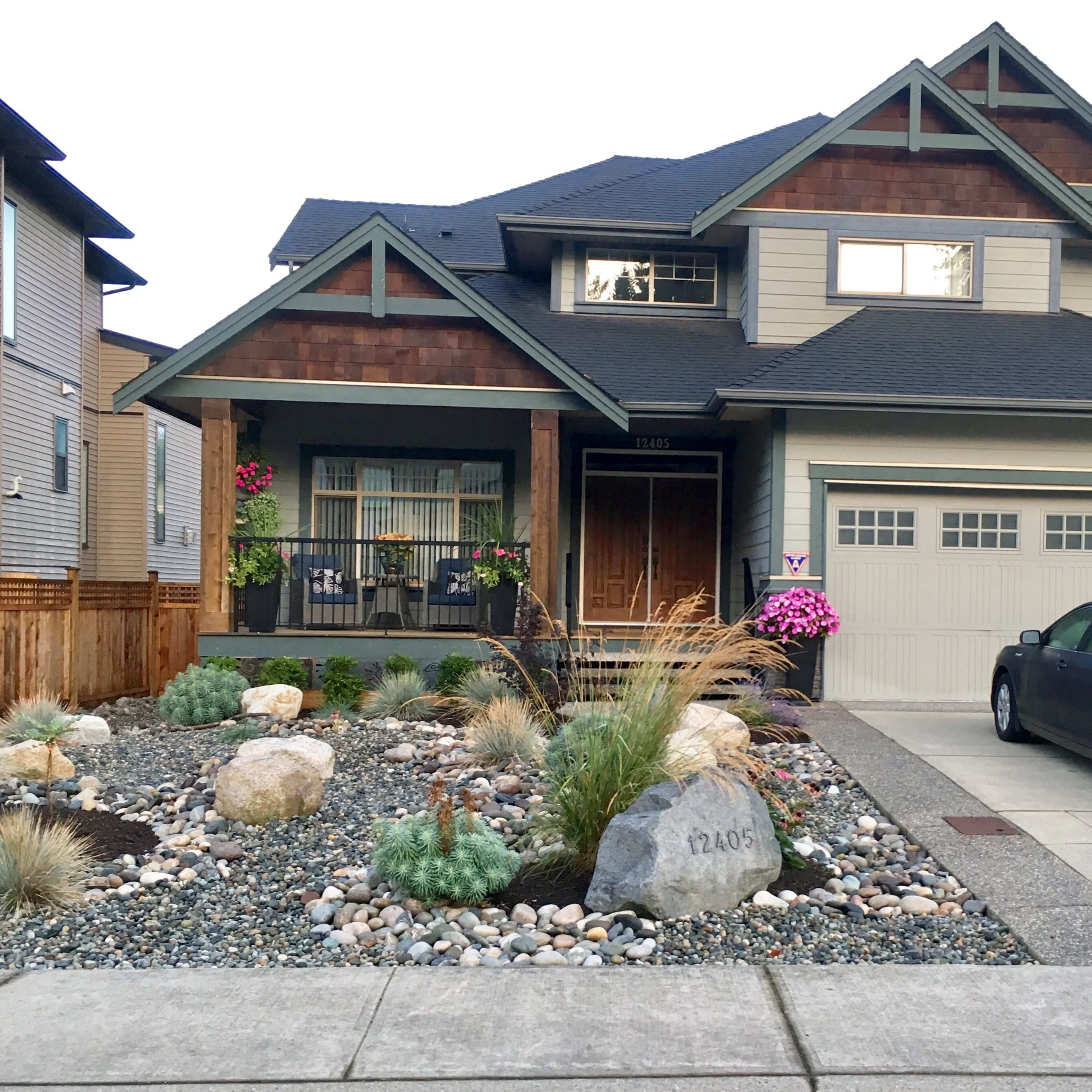 A two-story house with a mix of wood and siding features a landscaped front yard with rocks and plants, and a gray car parked in the driveway.