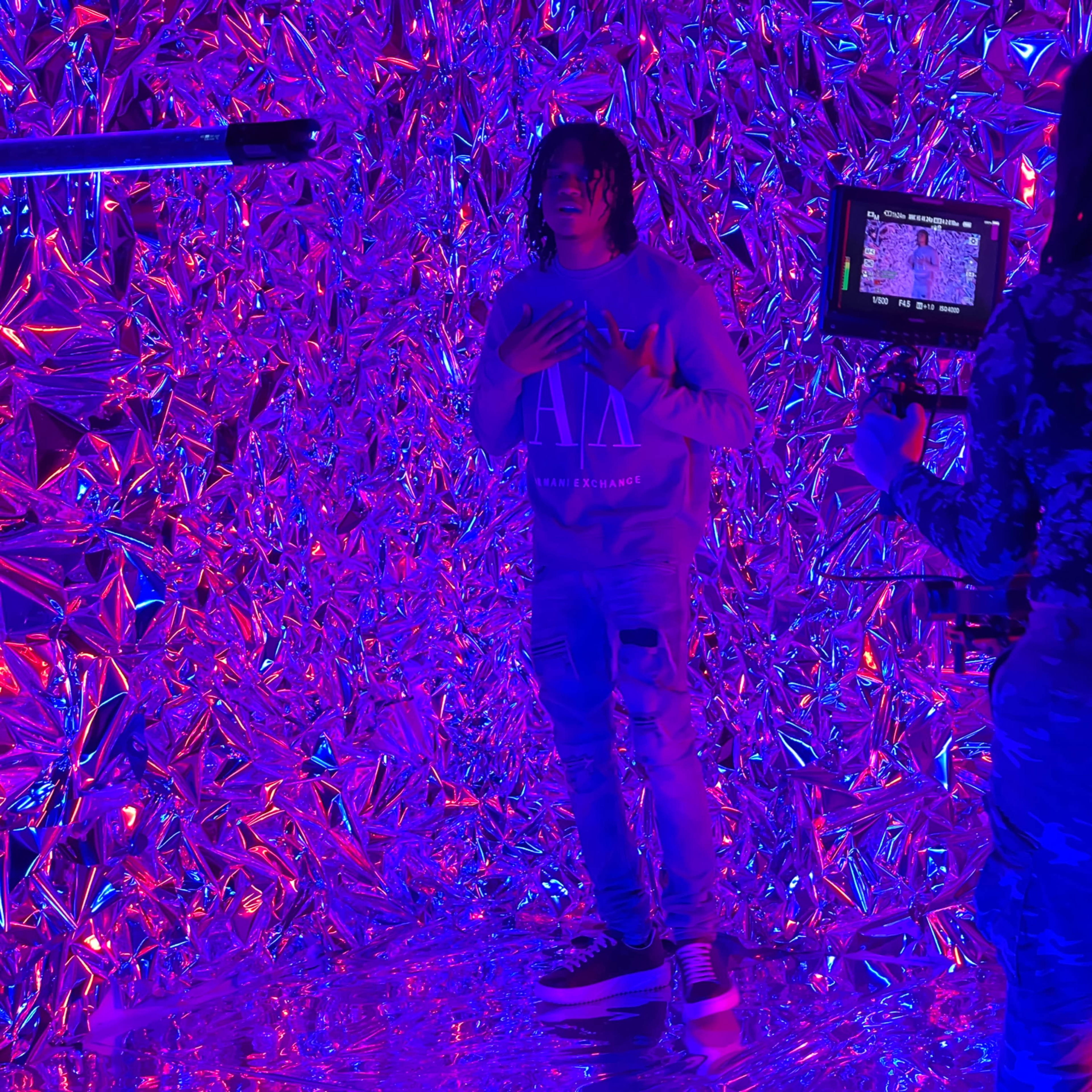 A person stands in front of a reflective, colorful background while another person films them with a camera.