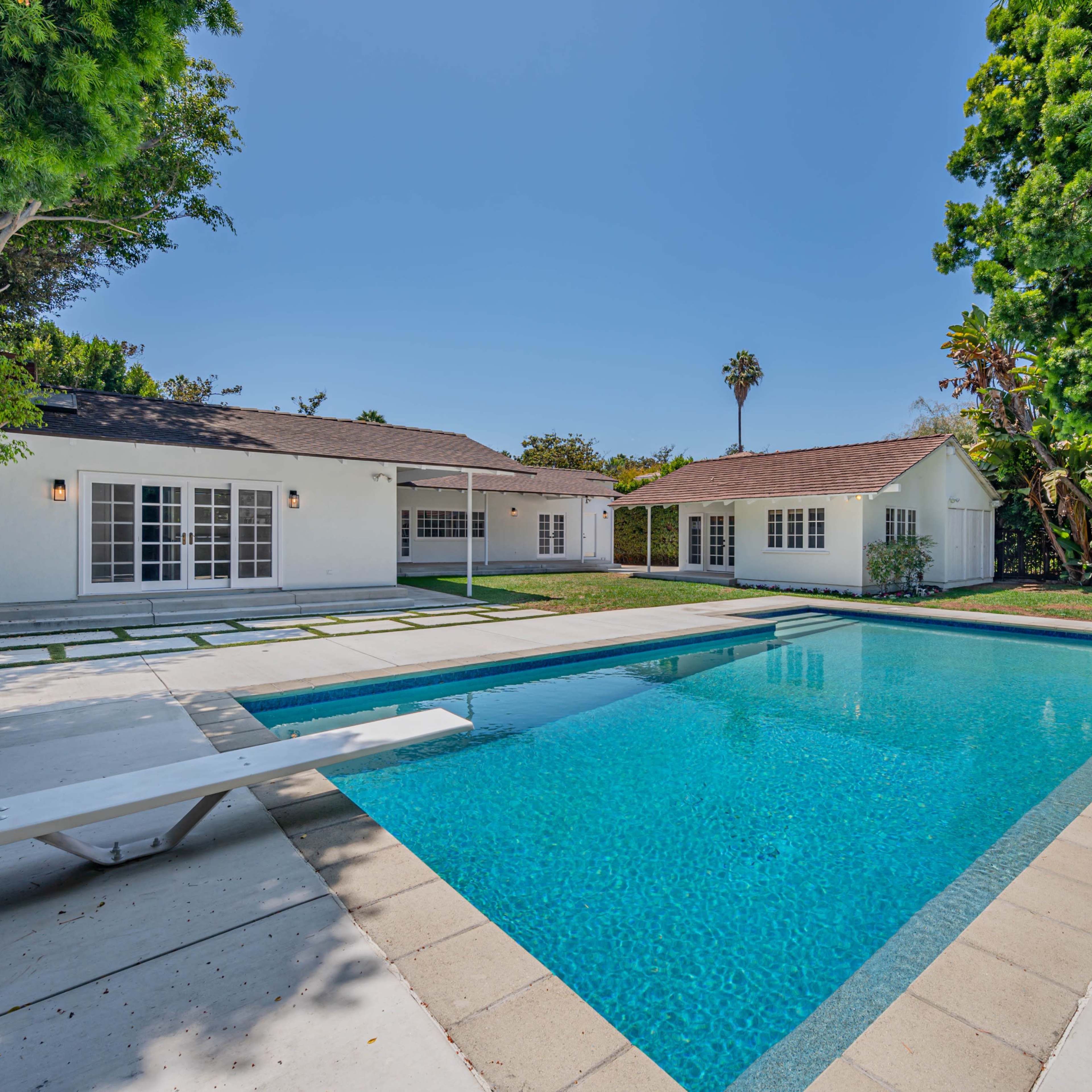 A backyard with a swimming pool surrounded by grass and trees, adjacent to a white single-story house.