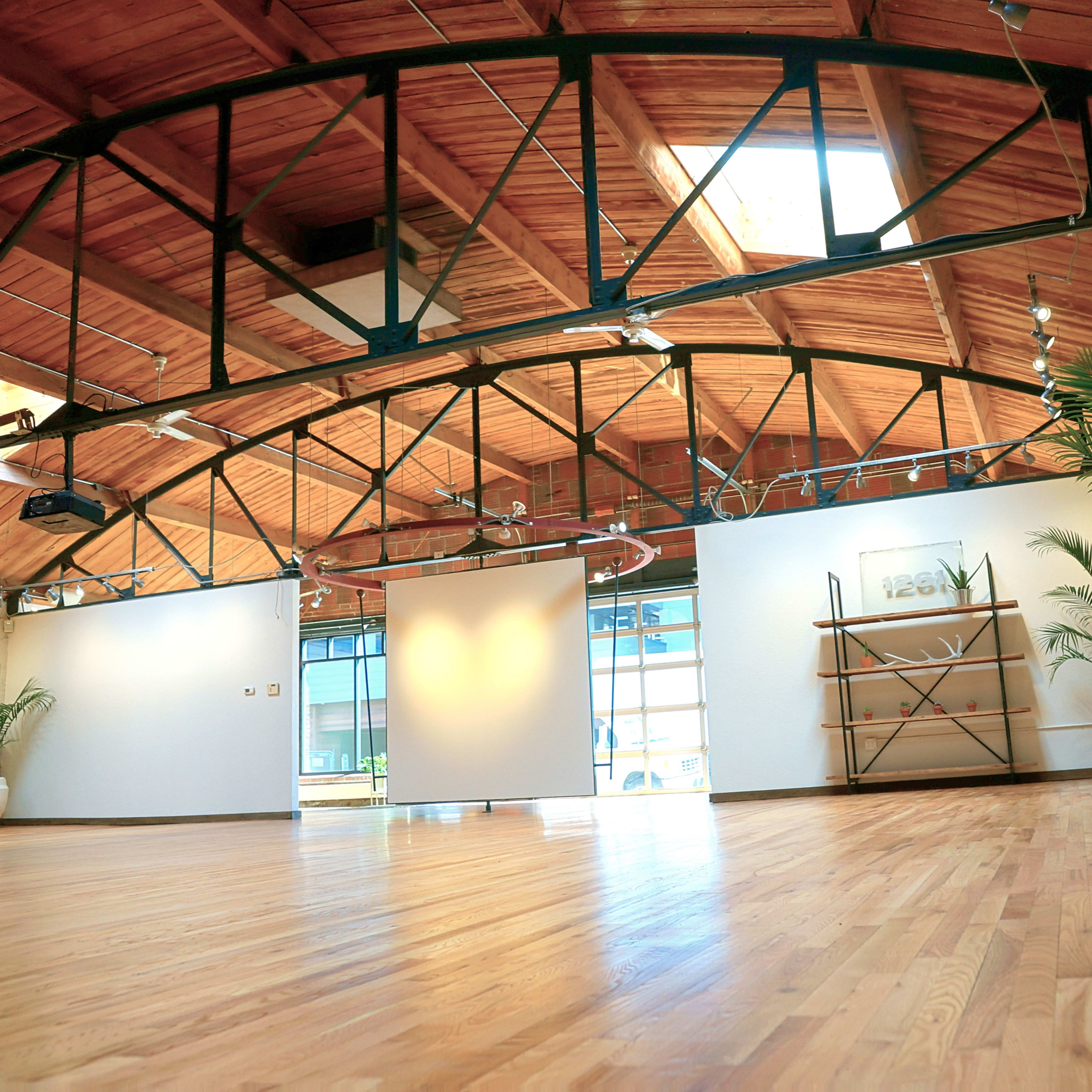 An empty studio space with wooden floors, large windows, and a high wooden-beamed ceiling, featuring potted plants and minimal furnishings.