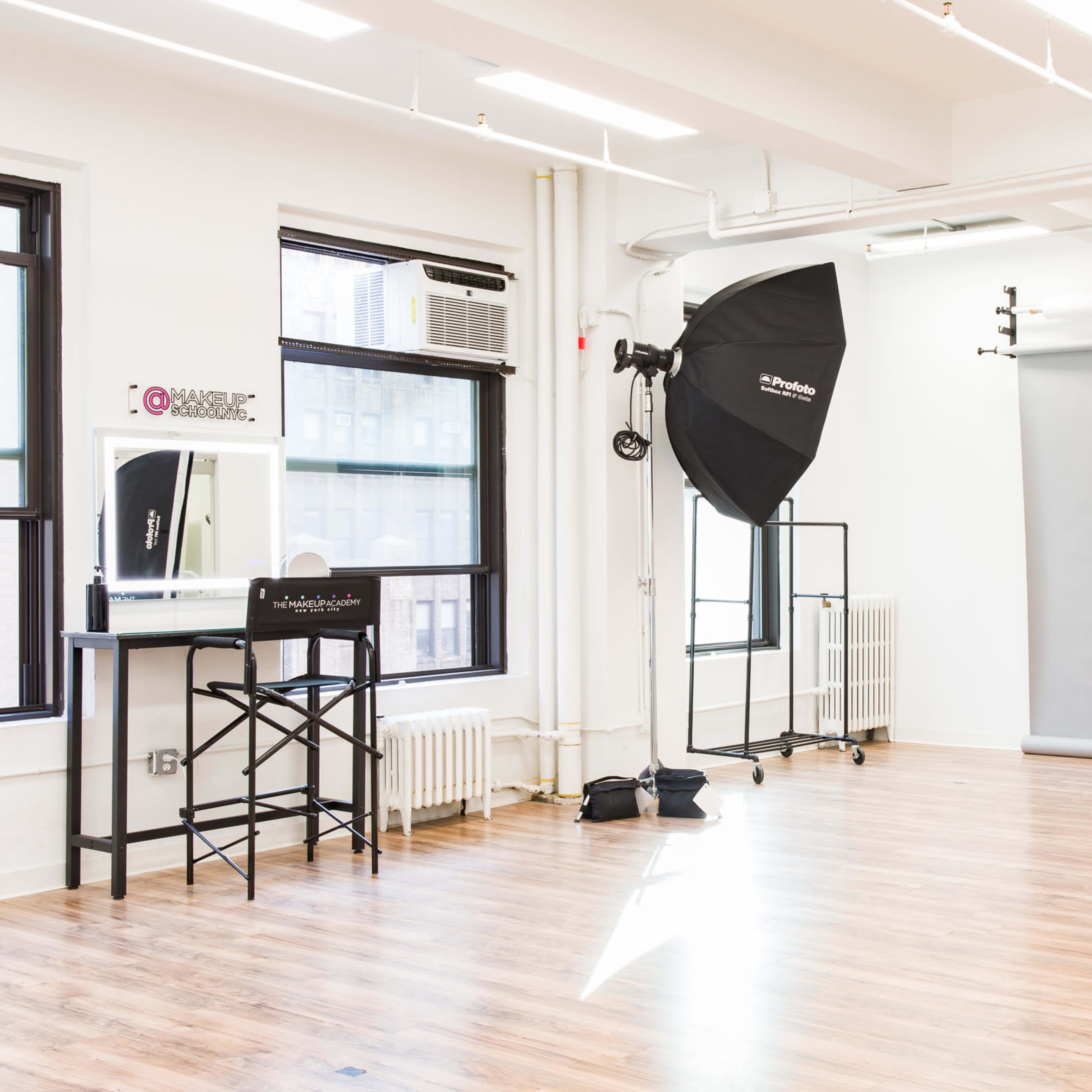 A photography studio with a large gray backdrop, two softboxes set up for lighting, and a makeup station with a mirror and chair.
