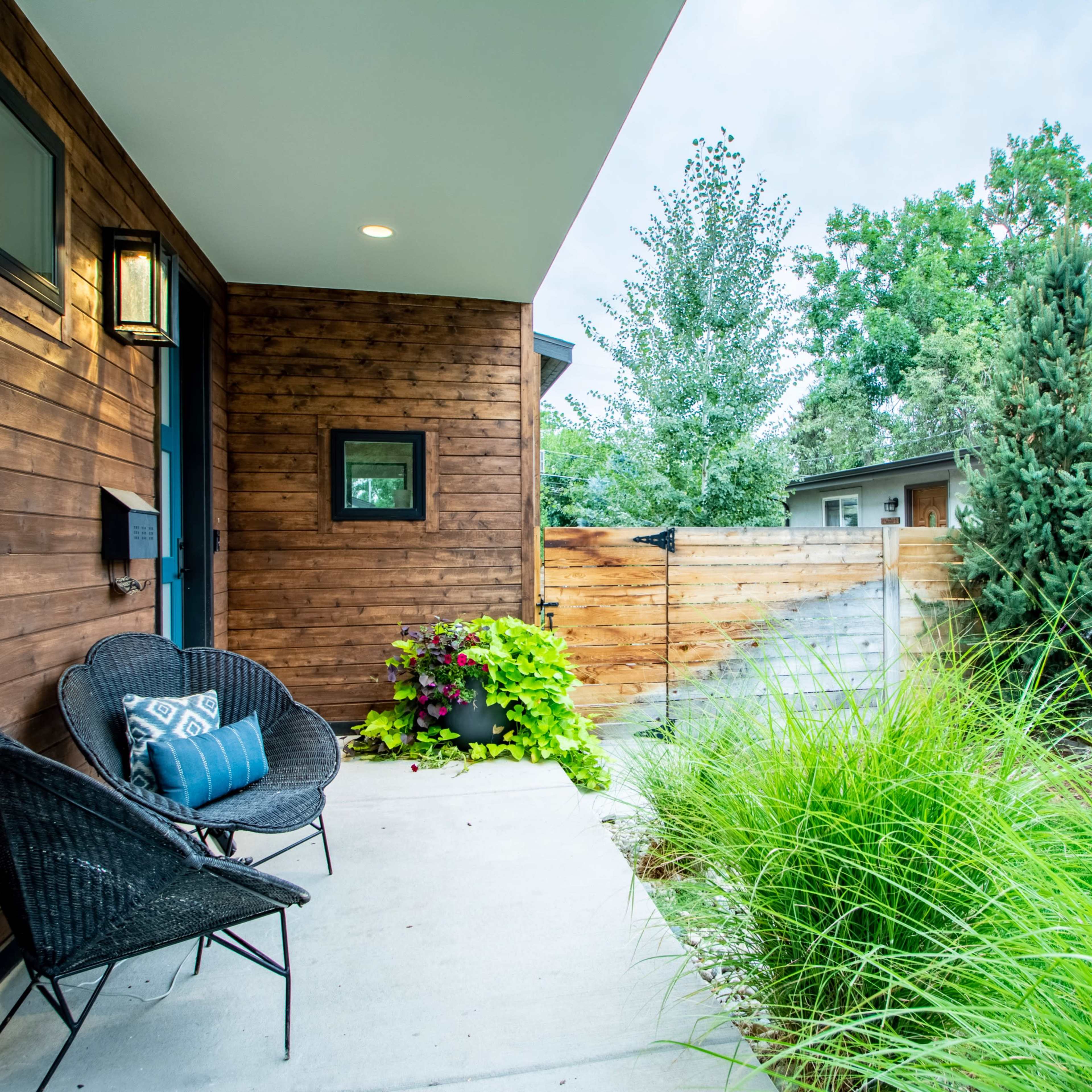 A porch area features two black chairs beside potted plants and tall grass, framed by wooden siding and trees.