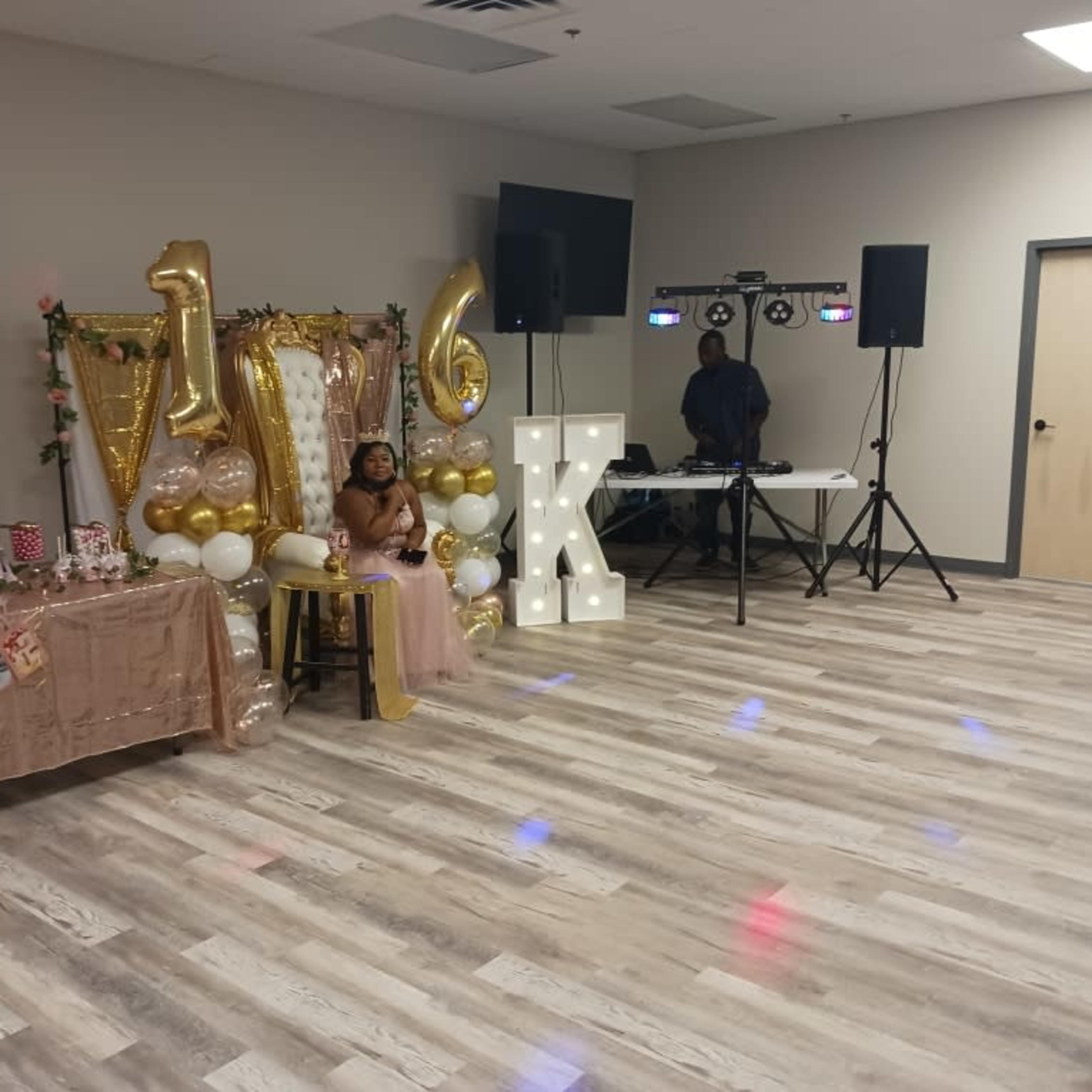 A child sits on a chair in front of a decorated table with a "16" sign and party supplies in a reception hall.