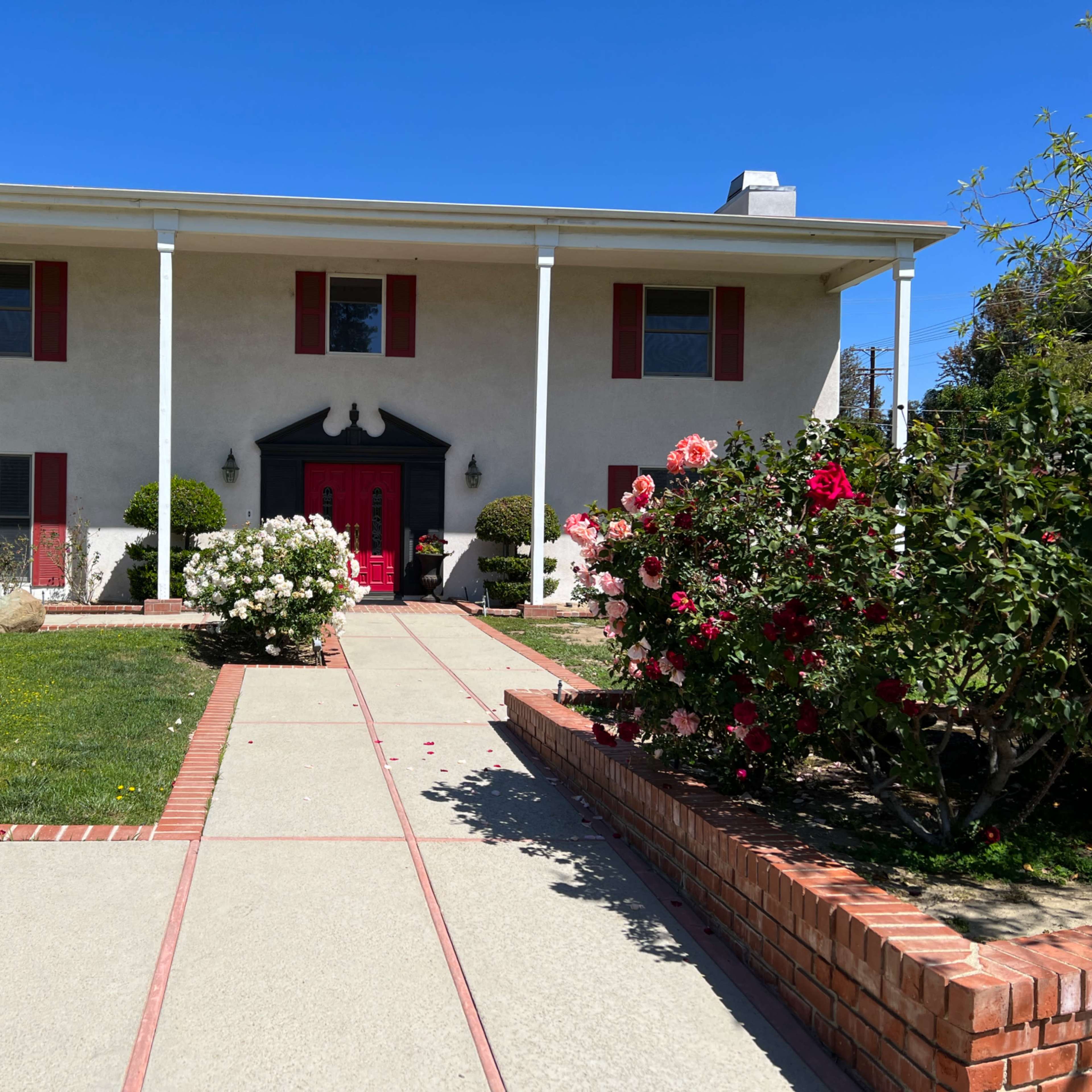 A two-story house with red shutters and a red front door, surrounded by flowering shrubs and a concrete pathway.