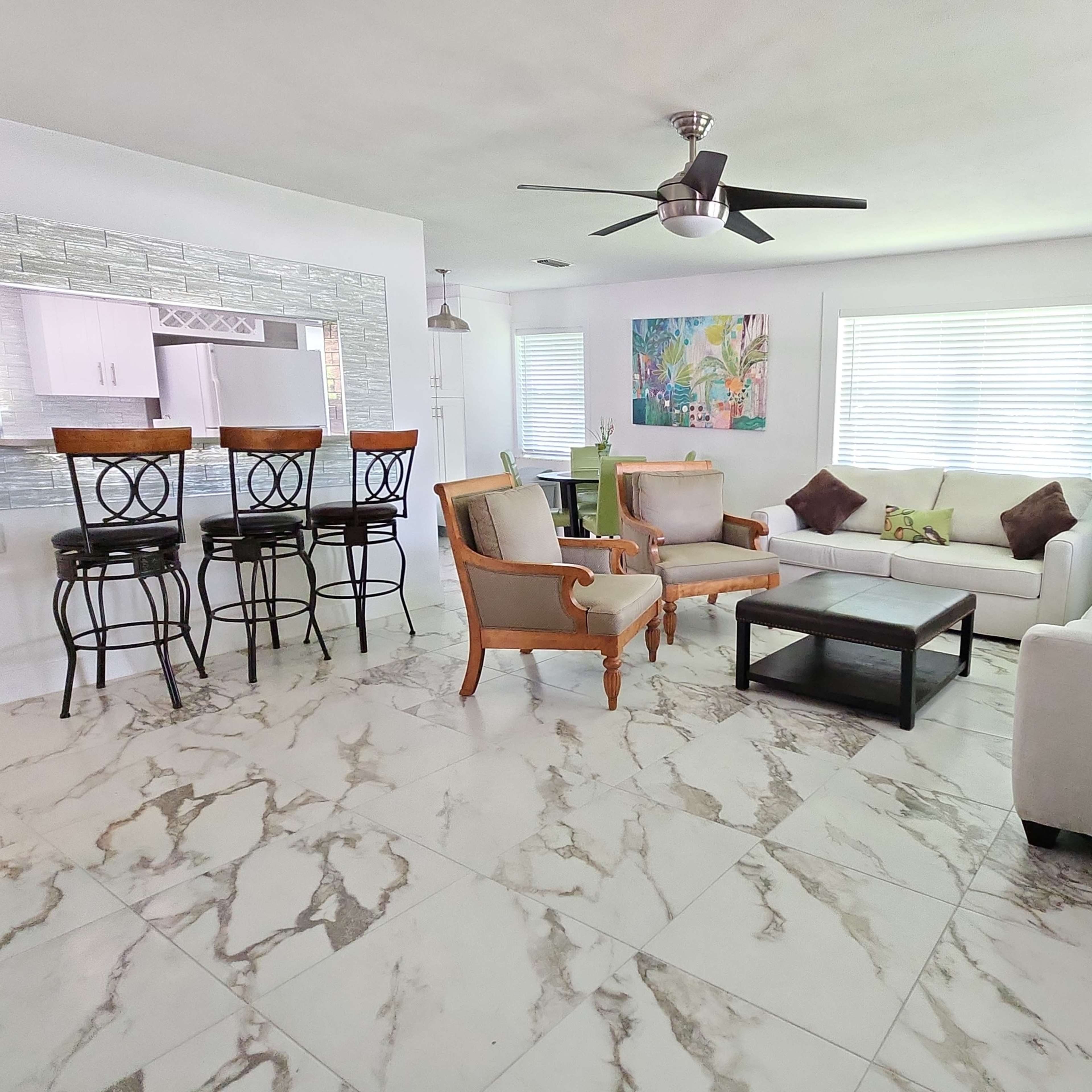 A modern living room with white furniture, marble-patterned flooring, and large windows providing natural light.