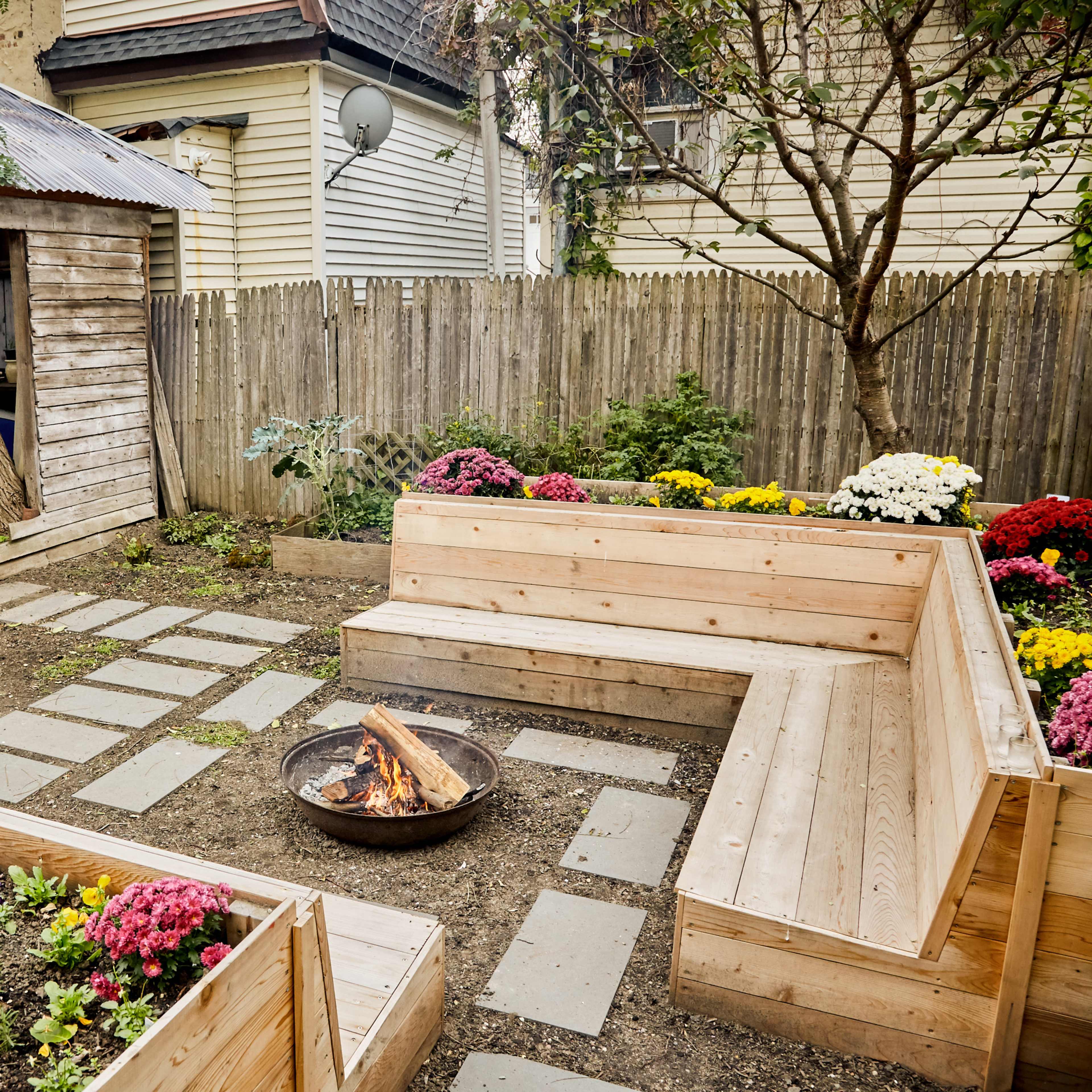 A cozy backyard with a wooden shed, a circular fire pit surrounded by wooden benches, and flower beds filled with colorful blooming plants.