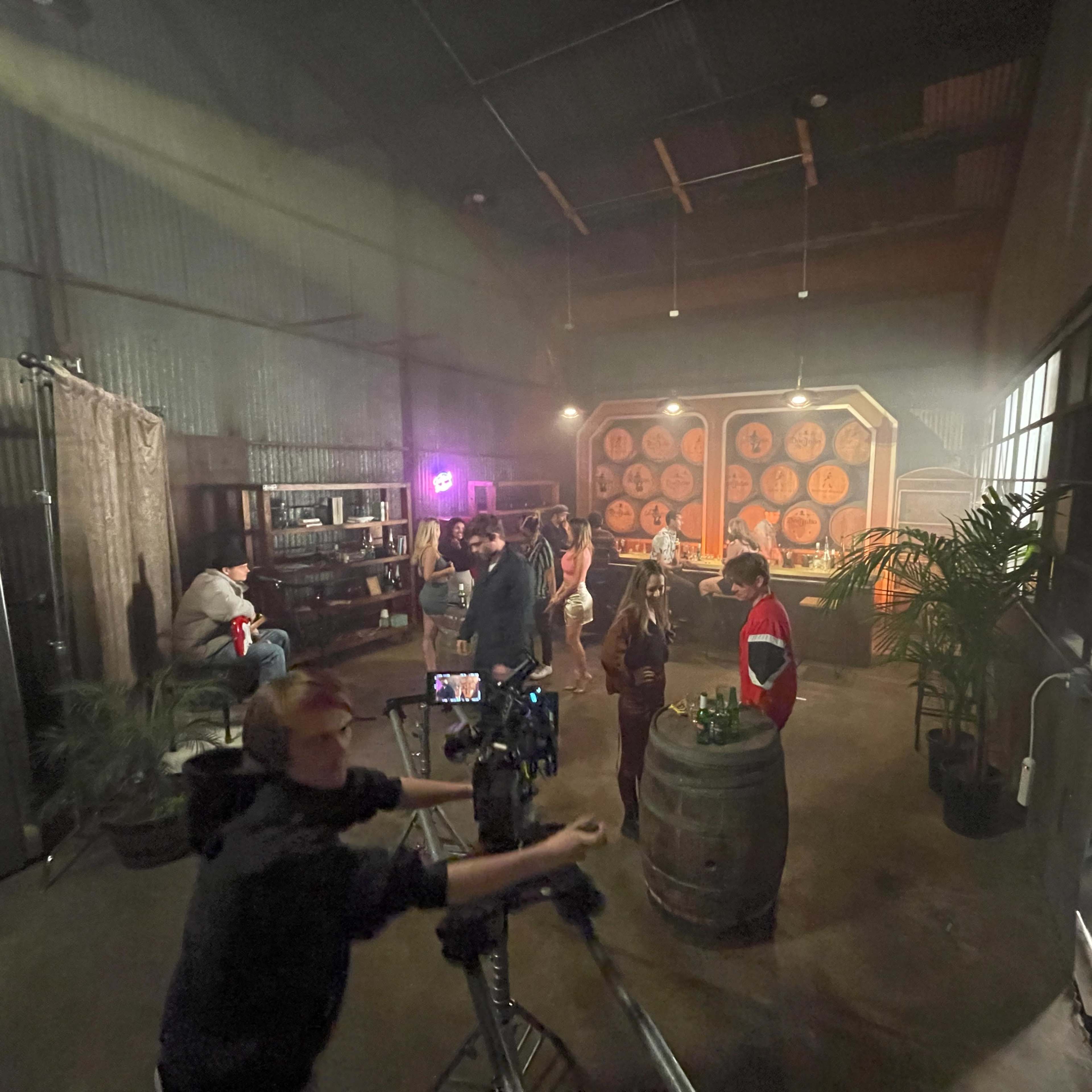 A group of people is gathered in a dimly lit warehouse setting with barrels and a backdrop of large circular decorations.