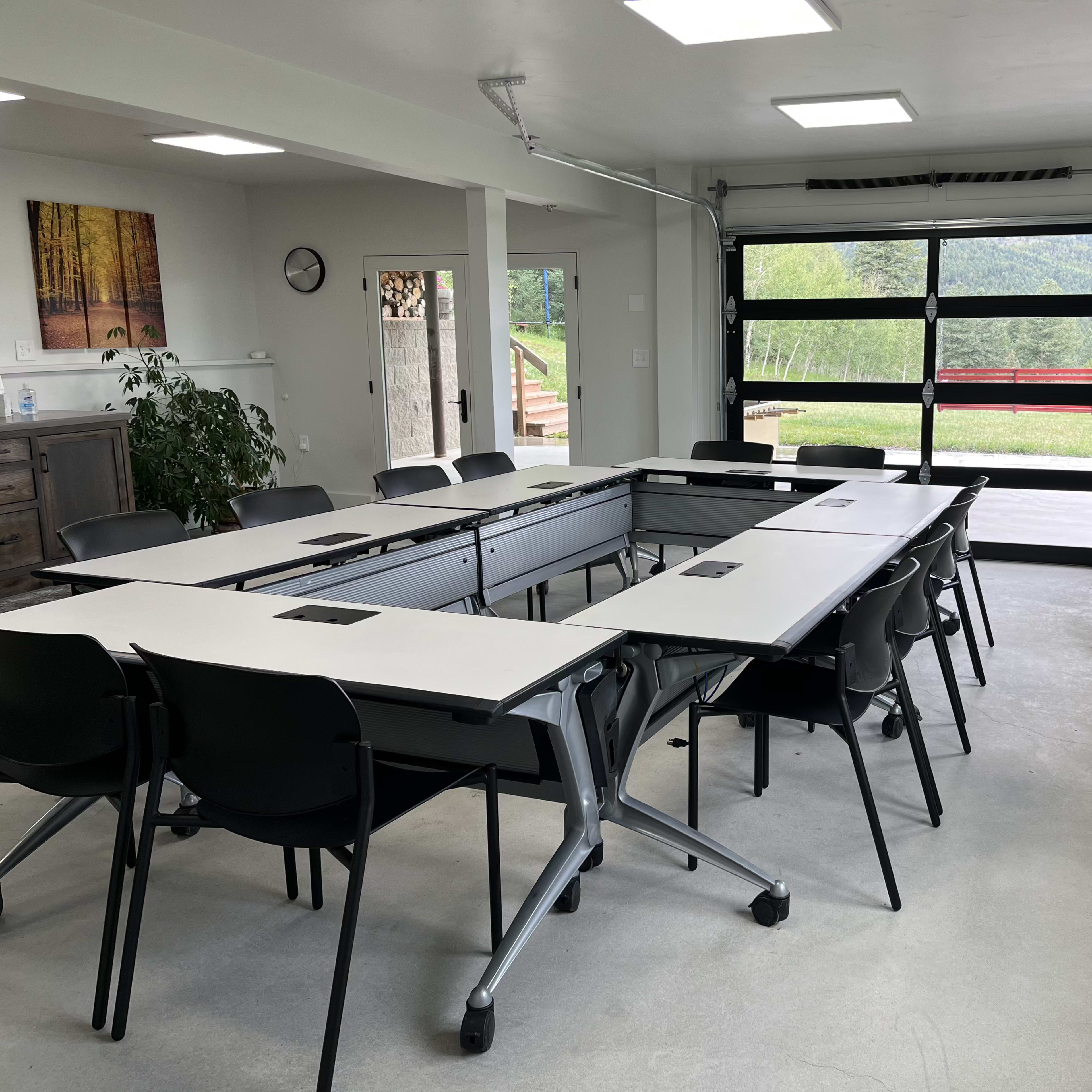 A modern conference room features a large table with movable sections and several black chairs, along with a glass door opening to a scenic view.