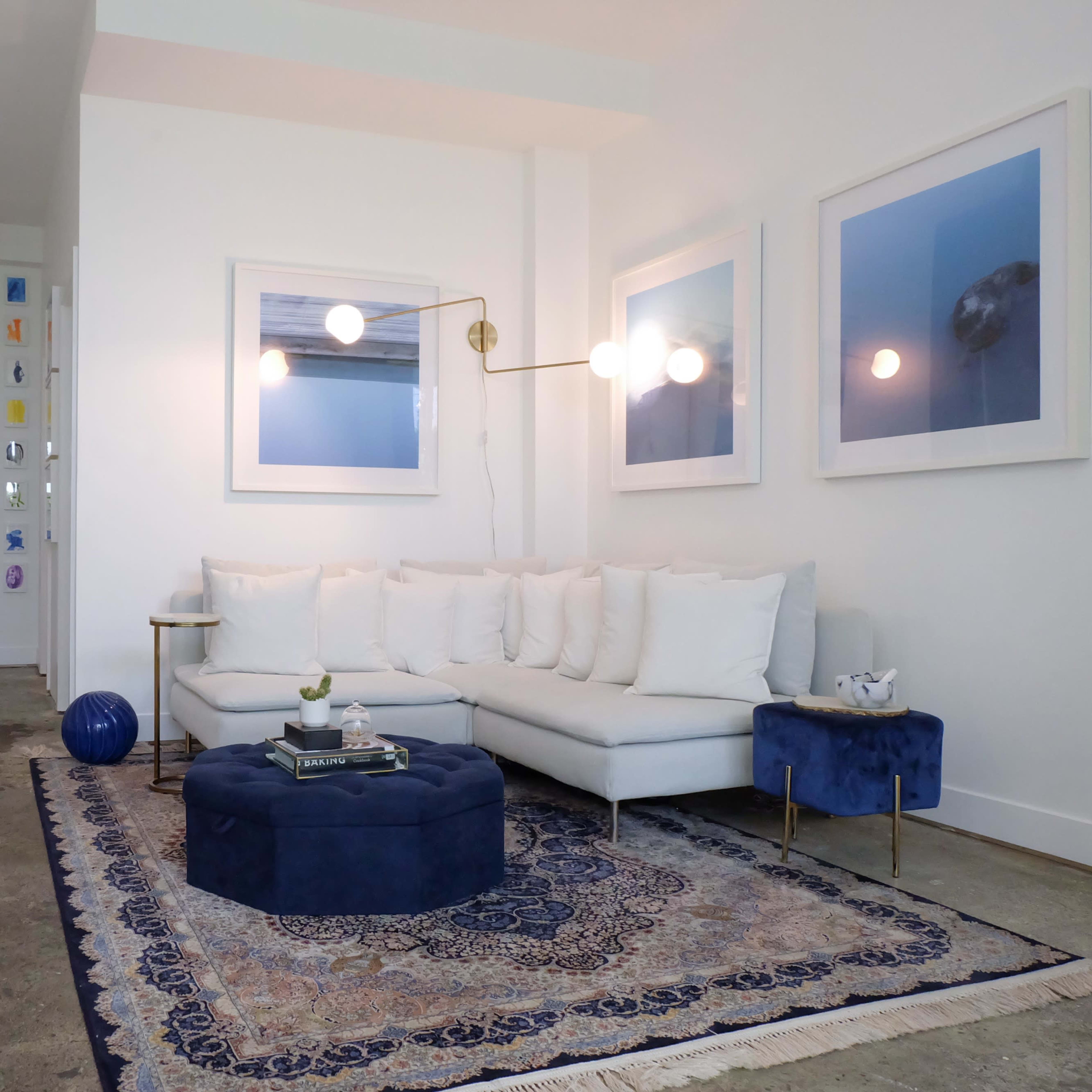A modern living room with a white sectional sofa, a round blue ottoman, and two framed artworks on the wall, all set against a light-colored, minimalist backdrop.