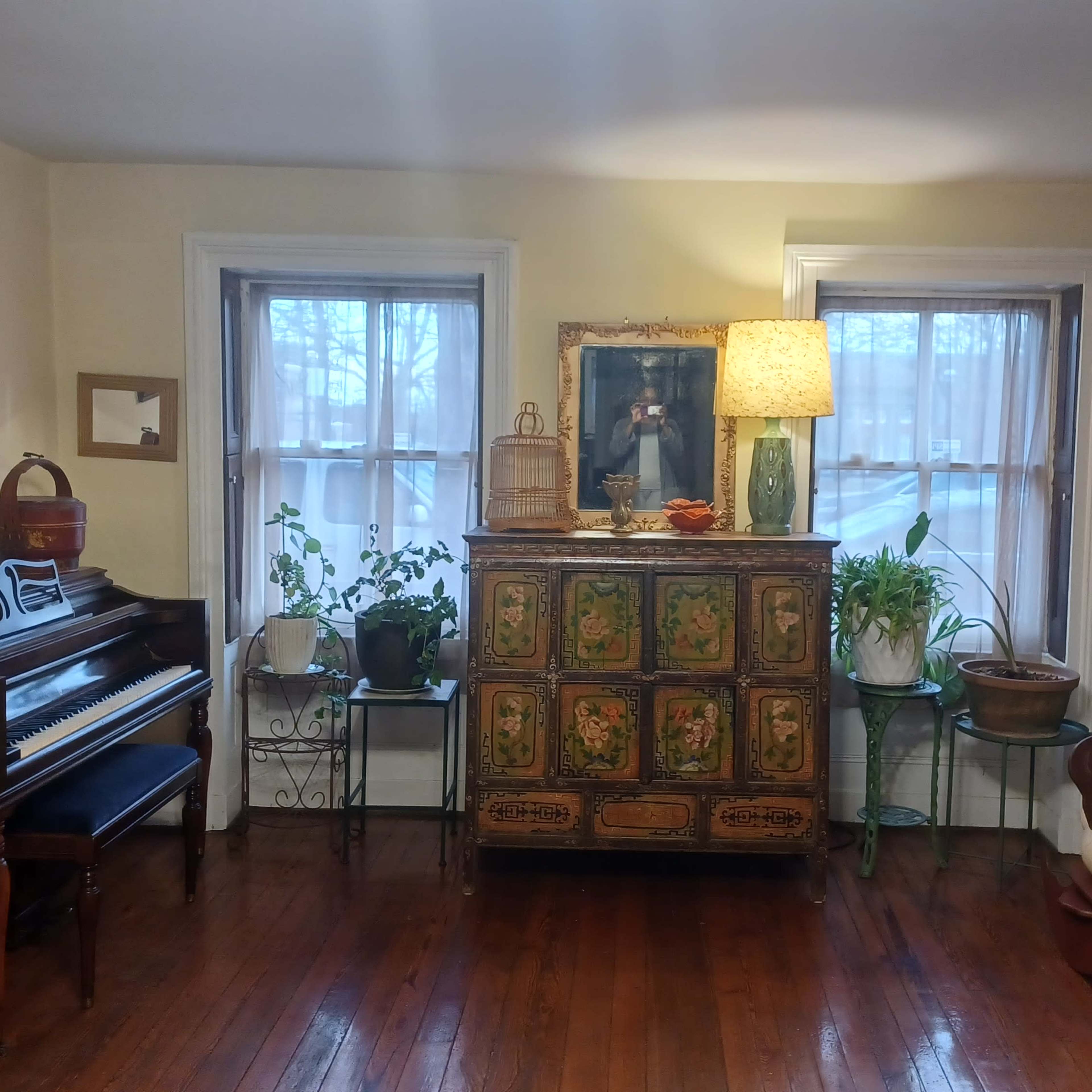A living room with a vintage piano, a floral-painted wooden cabinet, and various plants near two windows.