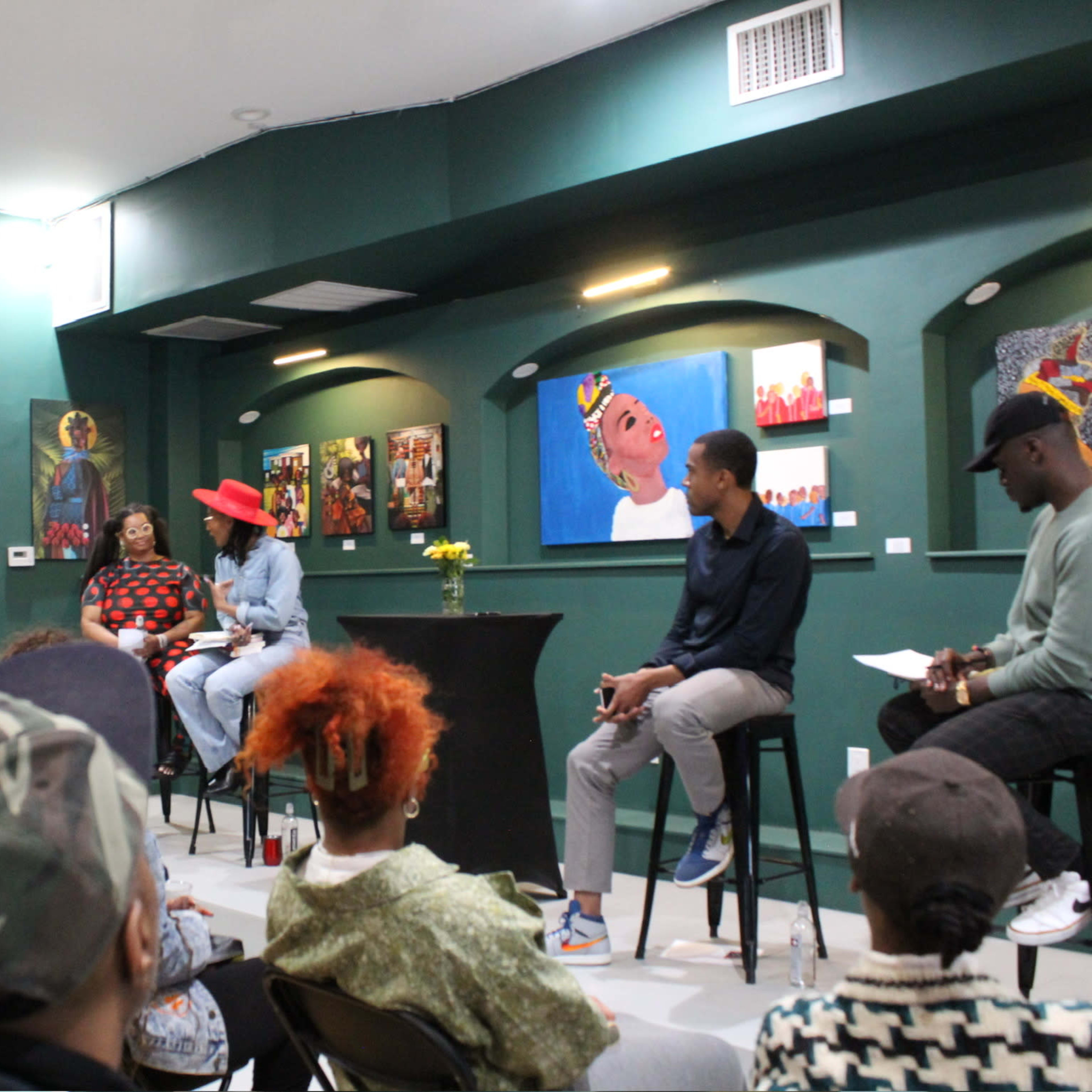 A group of people is seated in a small venue, attentively listening to a panel discussion featuring three speakers at a raised table.