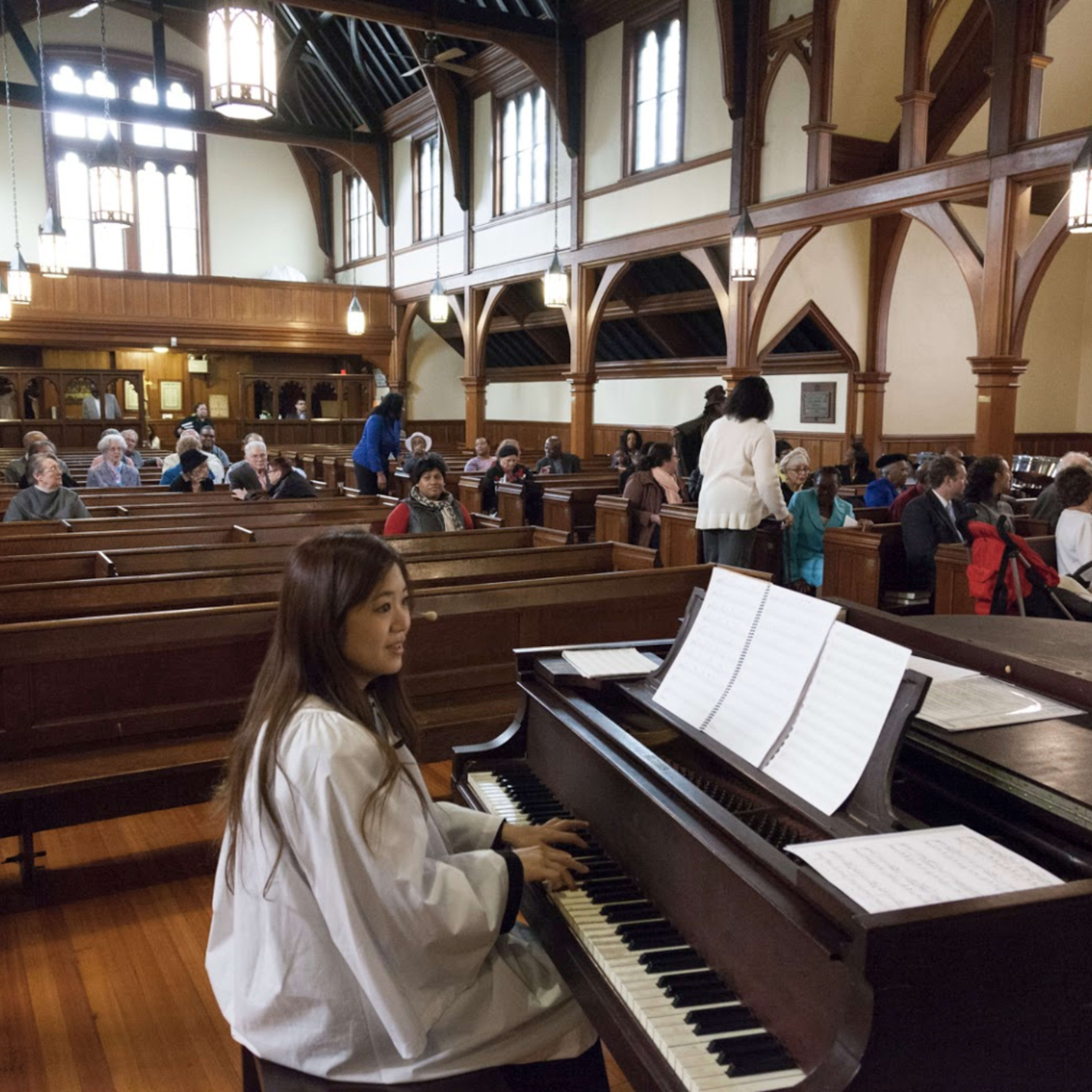 A pianist sits at a grand piano in a church, while a congregation gathers in the wooden pews for a service.