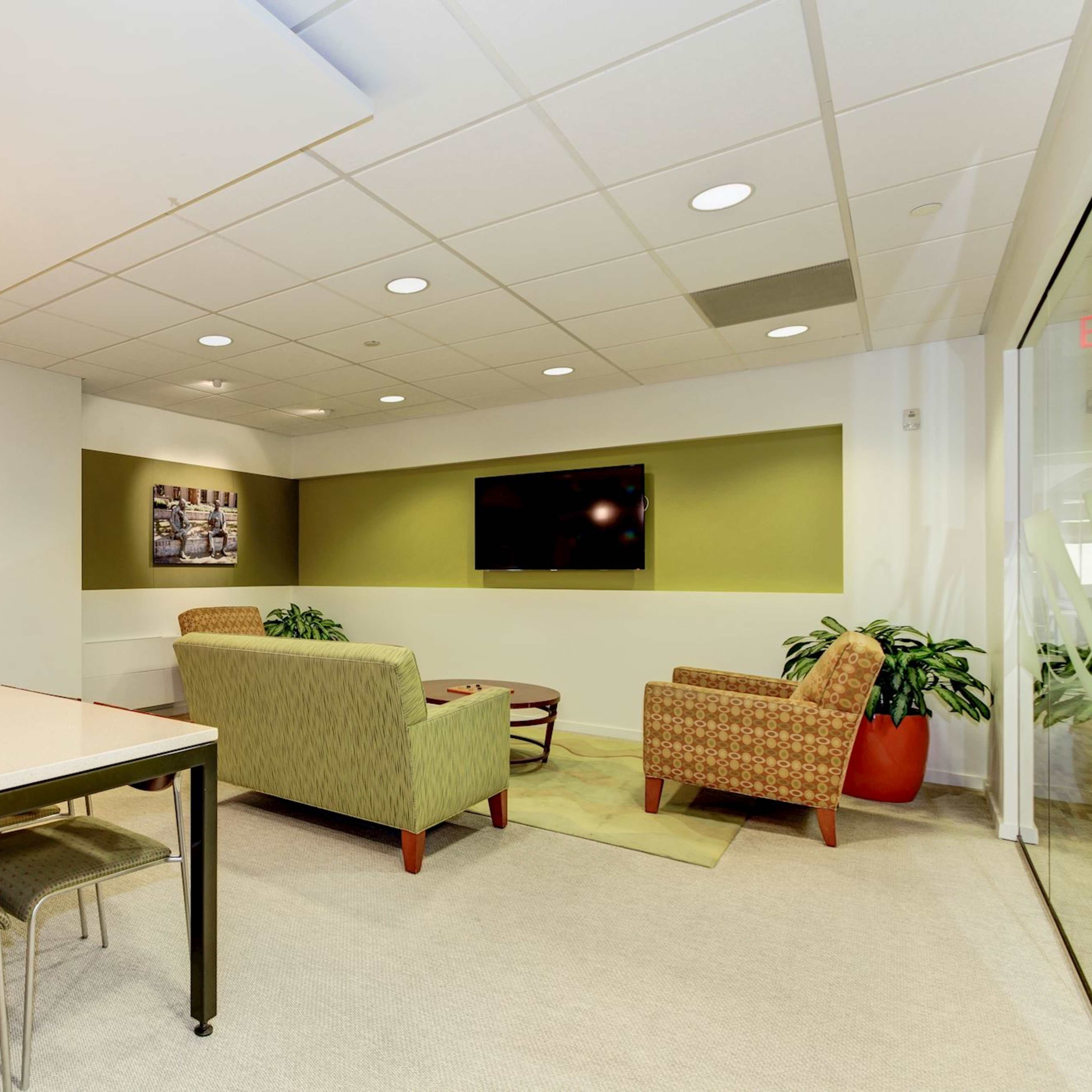 A modern meeting room featuring a large table with chairs, a seating area with a couch and armchair, and a TV mounted on the wall.