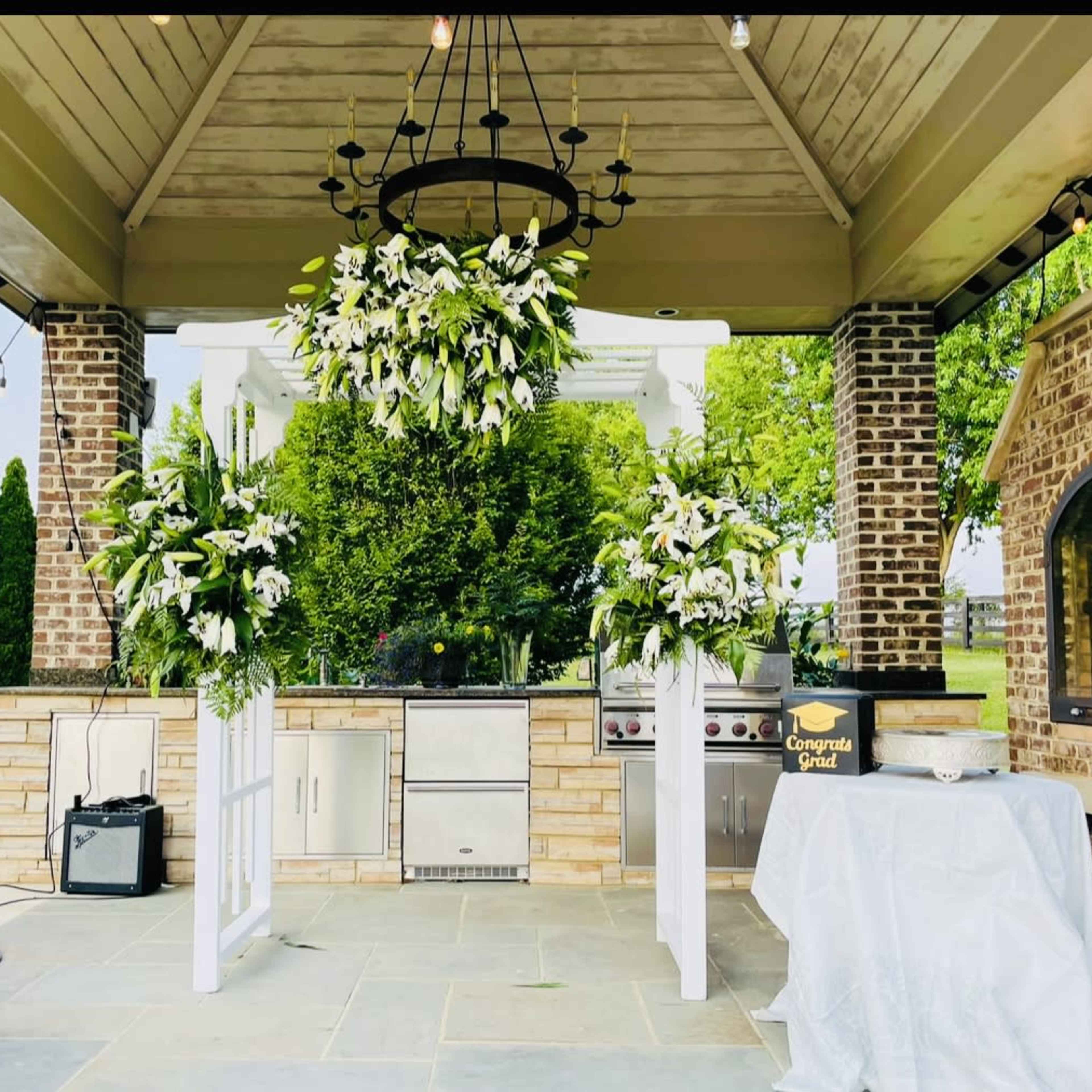 The outdoor kitchen area features a stone grill, a large floral arrangement hanging from the ceiling, and two side arrangements on pedestals, all set under a covered structure.