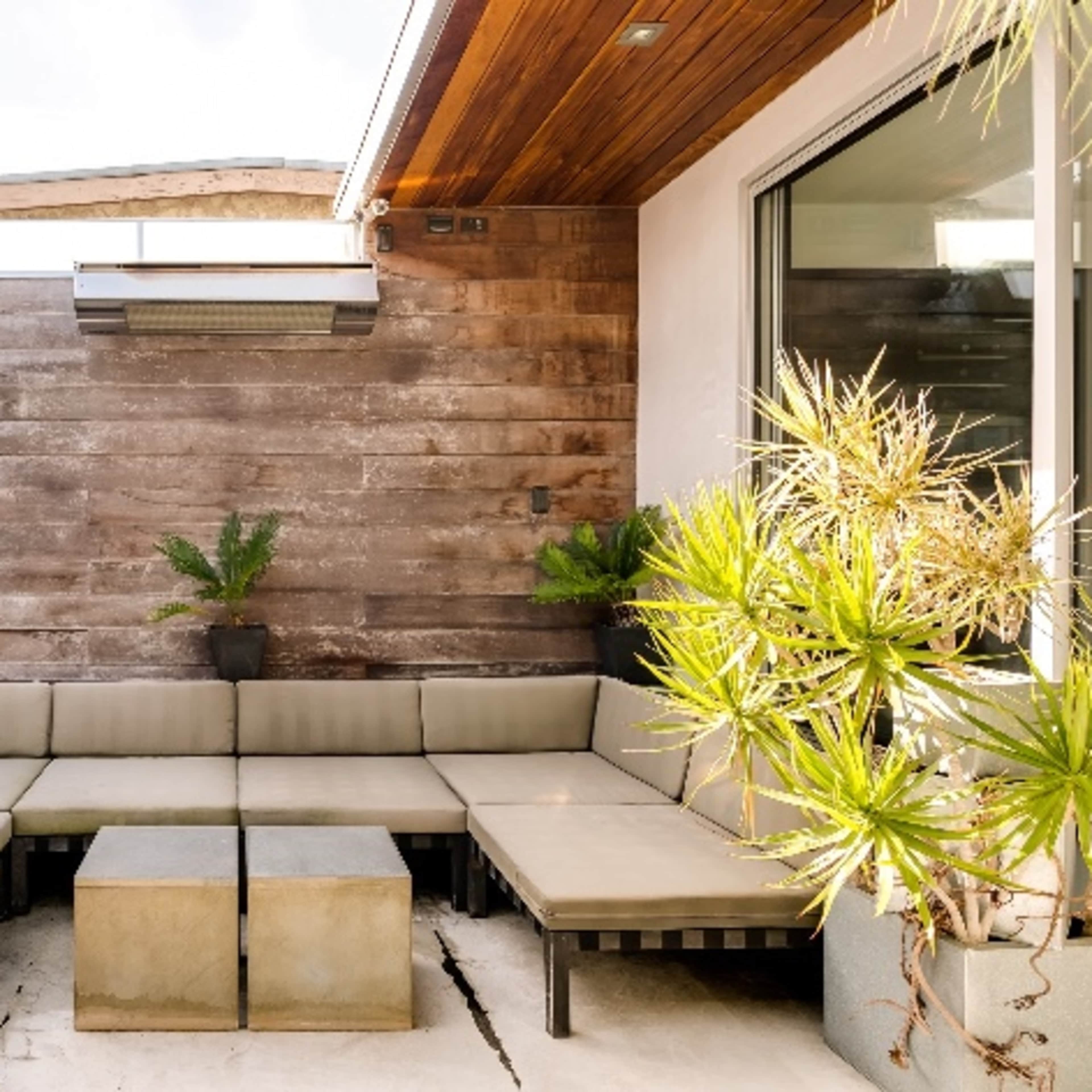 A modern outdoor seating area features a sectional sofa with beige cushions surrounding a concrete coffee table, complemented by potted plants against a wooden wall.