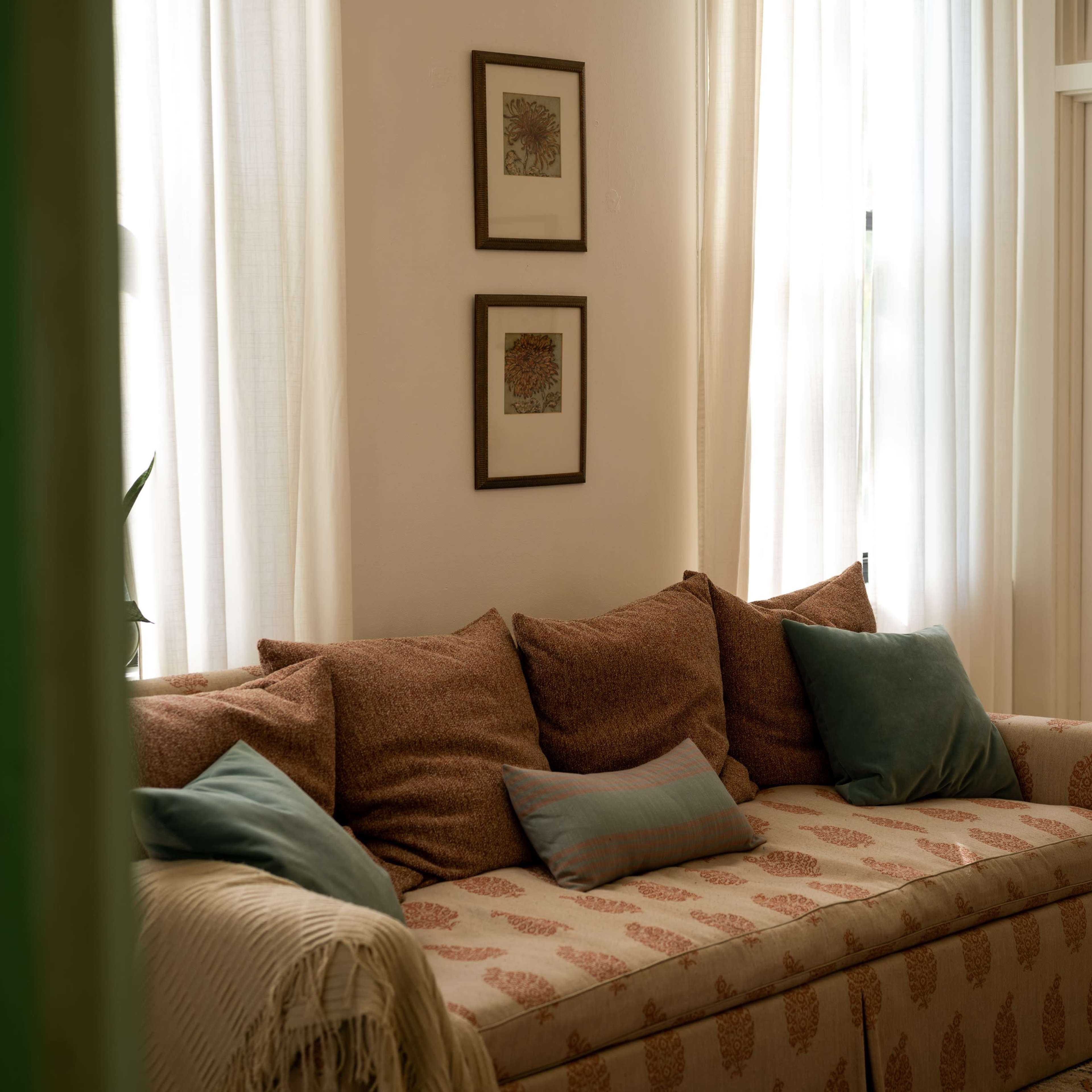 A patterned sofa with several cushions is positioned in a well-lit room featuring sheer curtains and framed artwork on the wall.