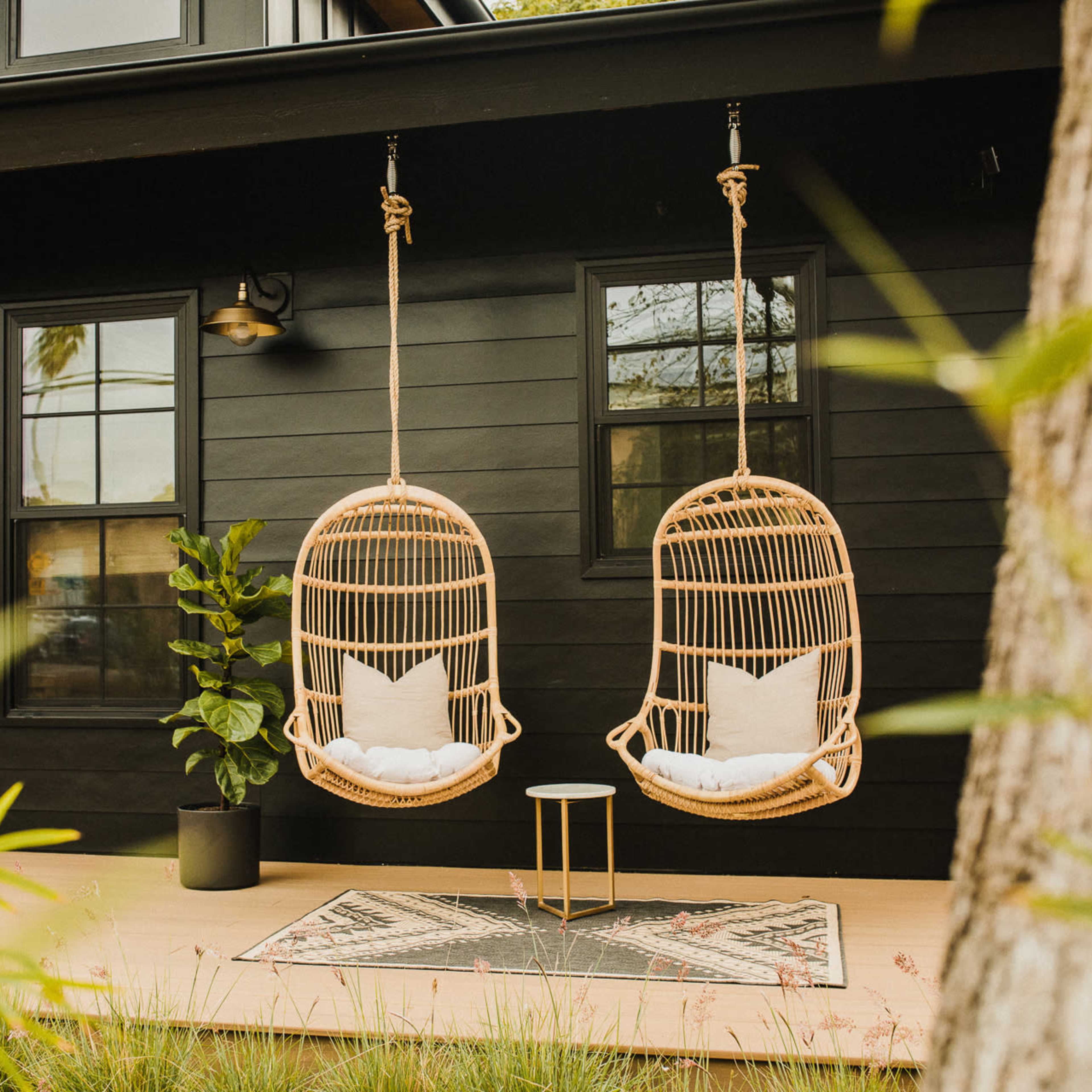 Two suspended rattan chairs with white cushions hang in front of a black house, accompanied by a small table and a decorative rug.