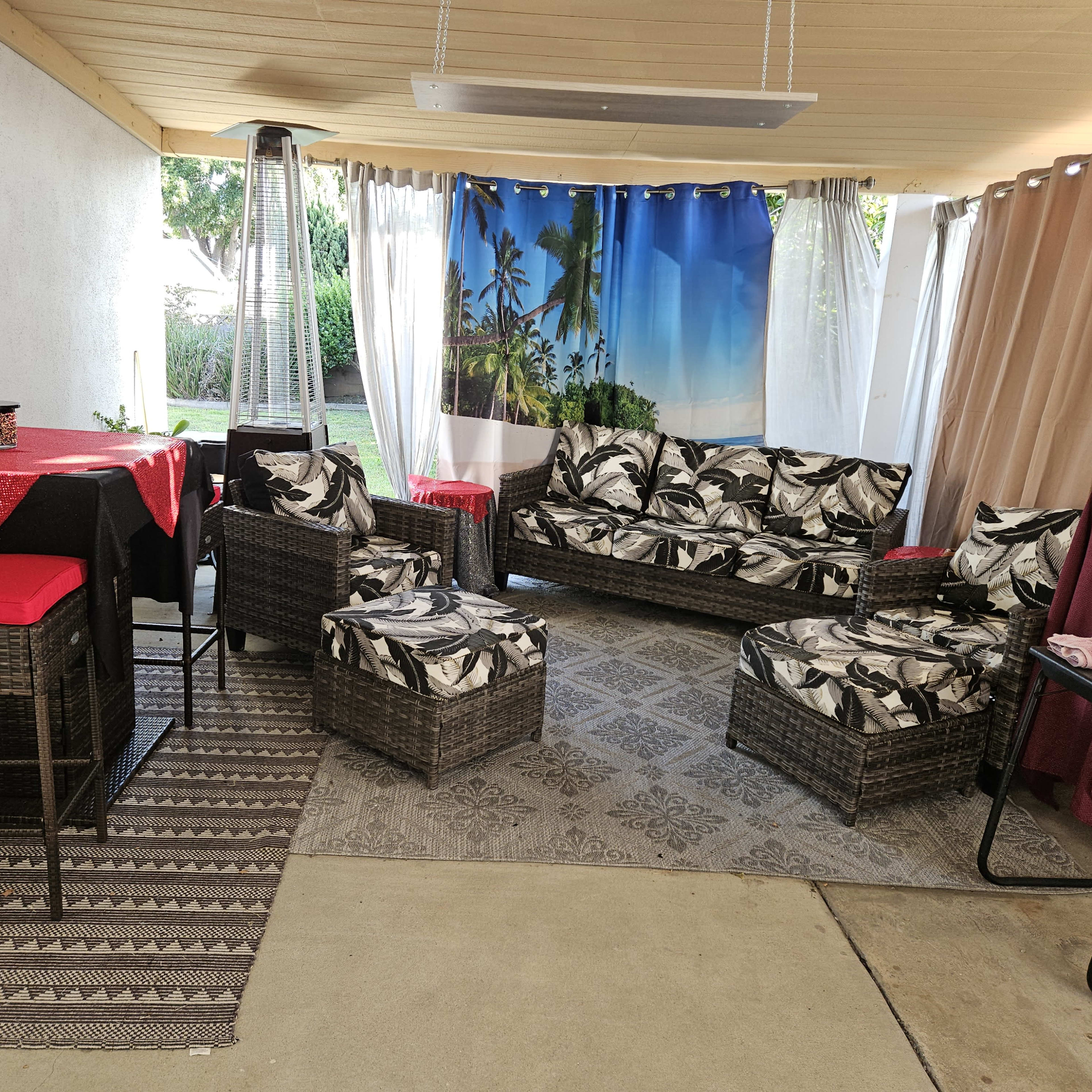 A covered outdoor seating area with wicker furniture, a red table, and a backdrop featuring palm trees and a blue sky.