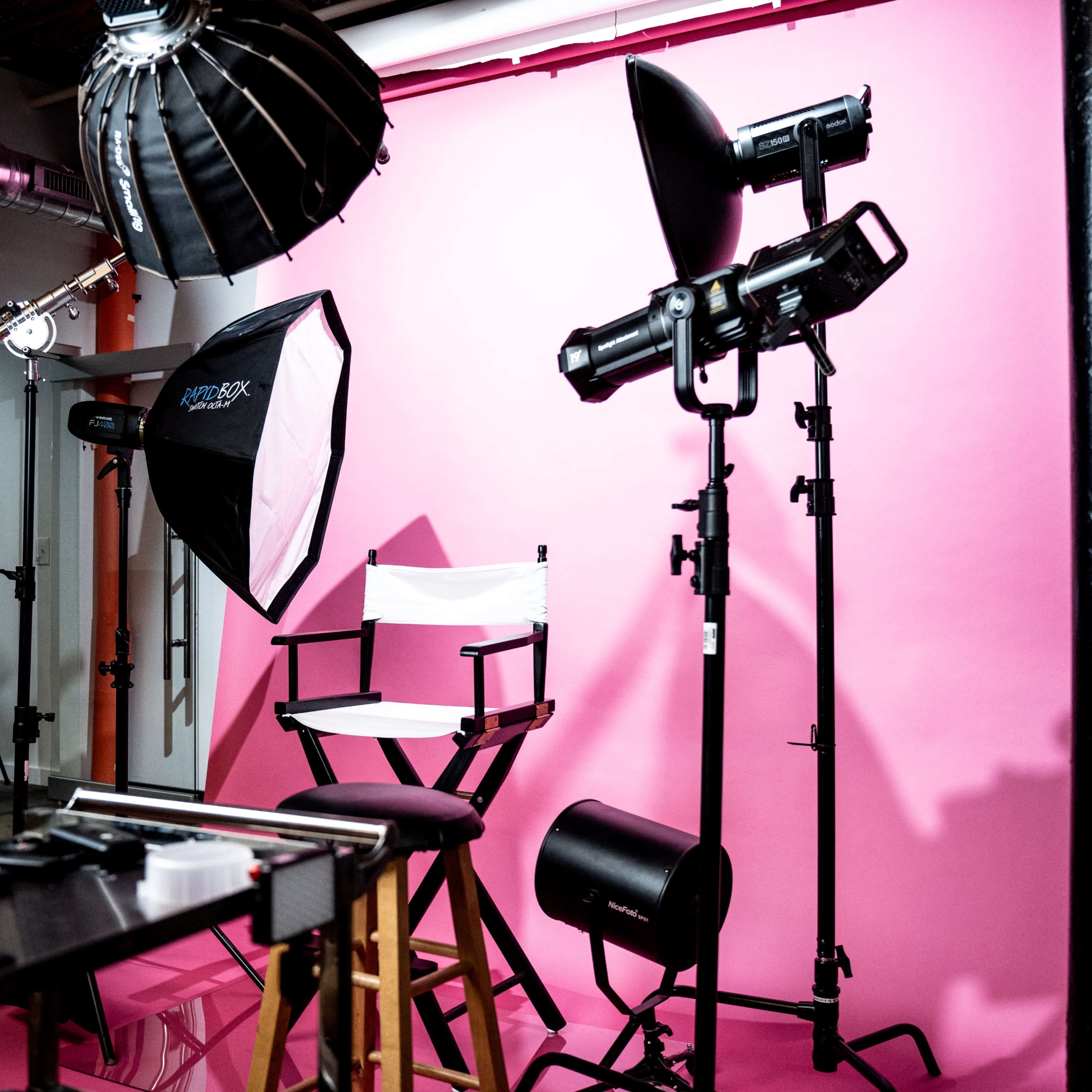 A photography studio setup features a pink background, various lighting equipment, and a director's chair.