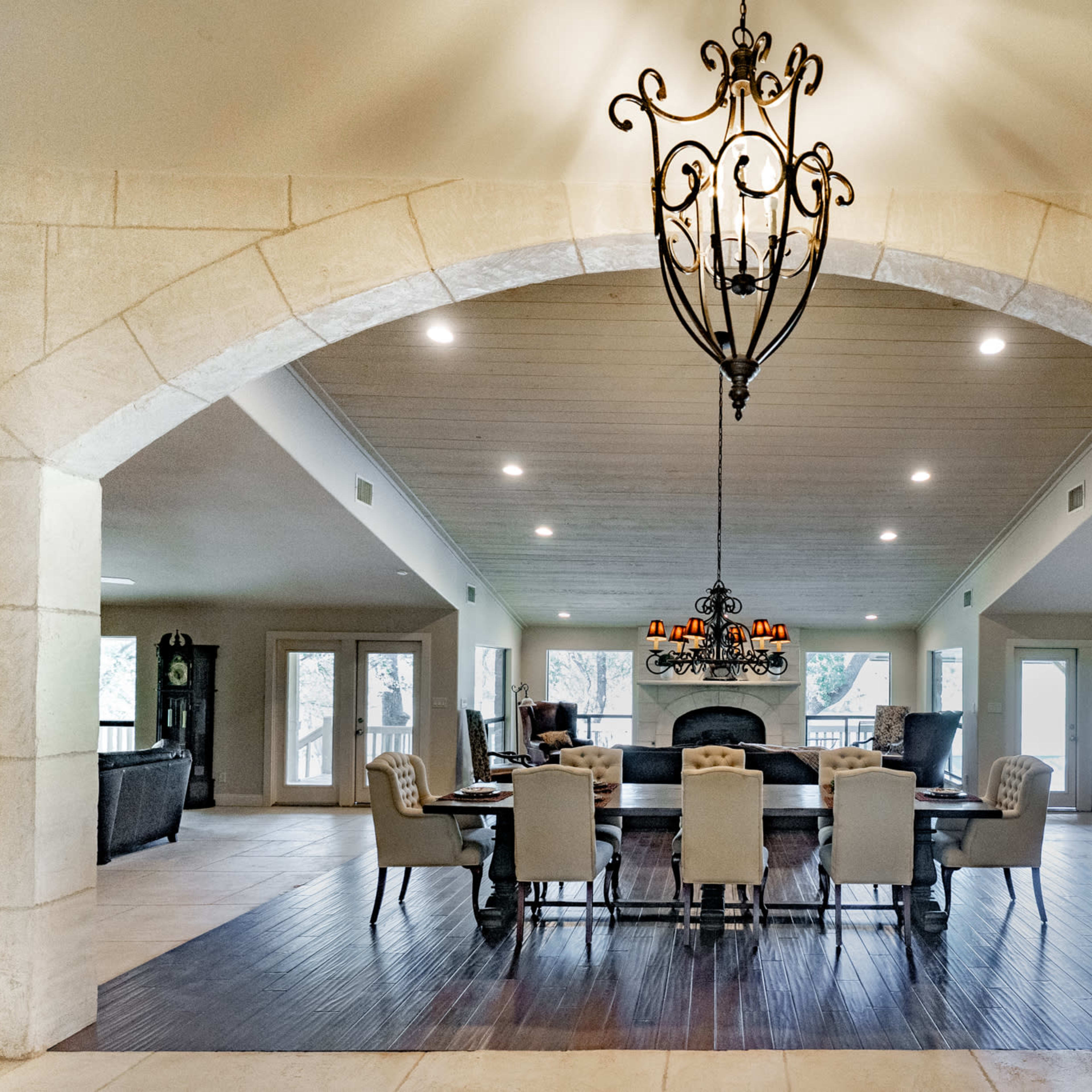 A spacious dining area featuring a long table surrounded by upholstered chairs, beneath an ornate chandelier, set within an arched stone entryway.