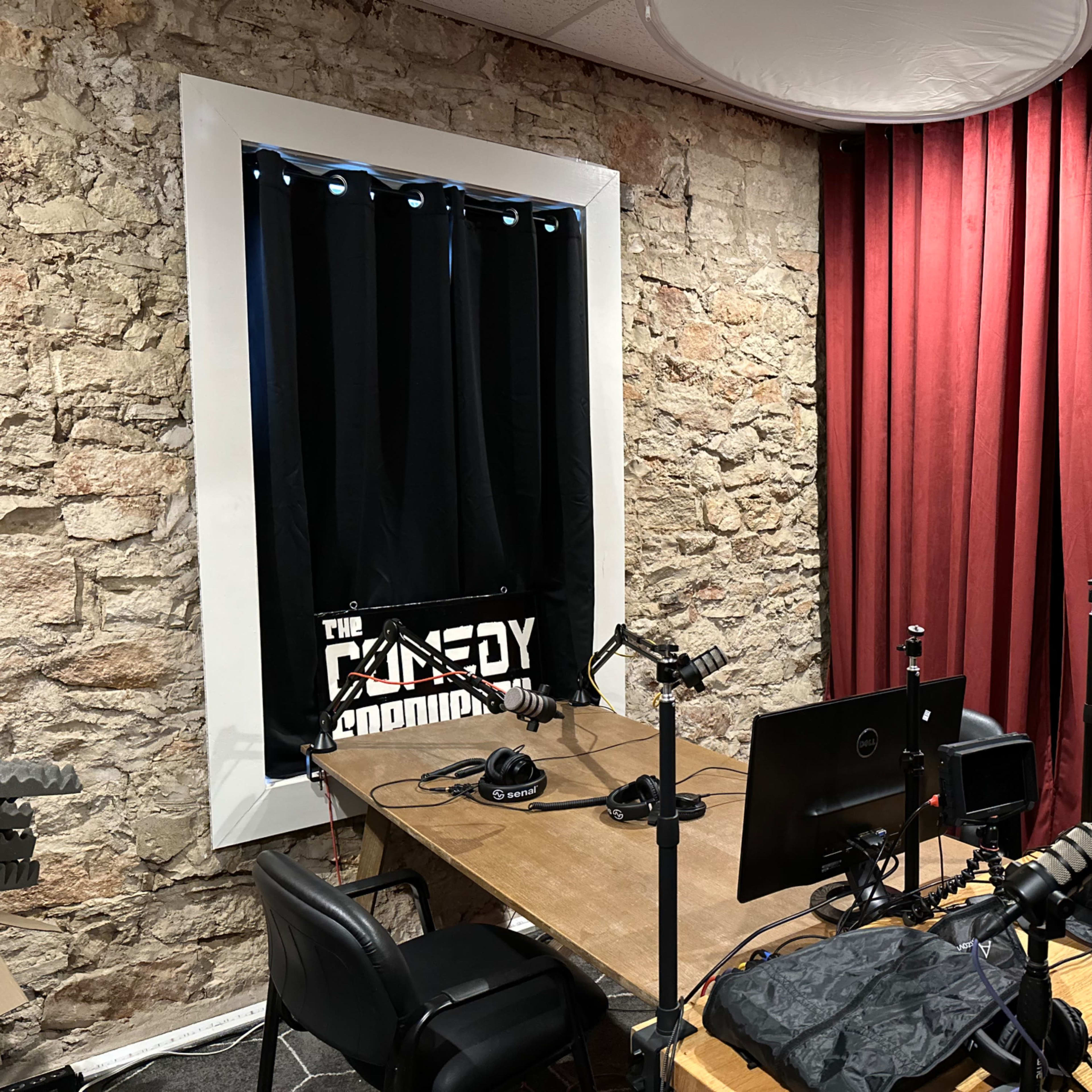 A recording studio with a wooden table, several microphones on stands, and a backdrop of red curtains next to a stone wall.