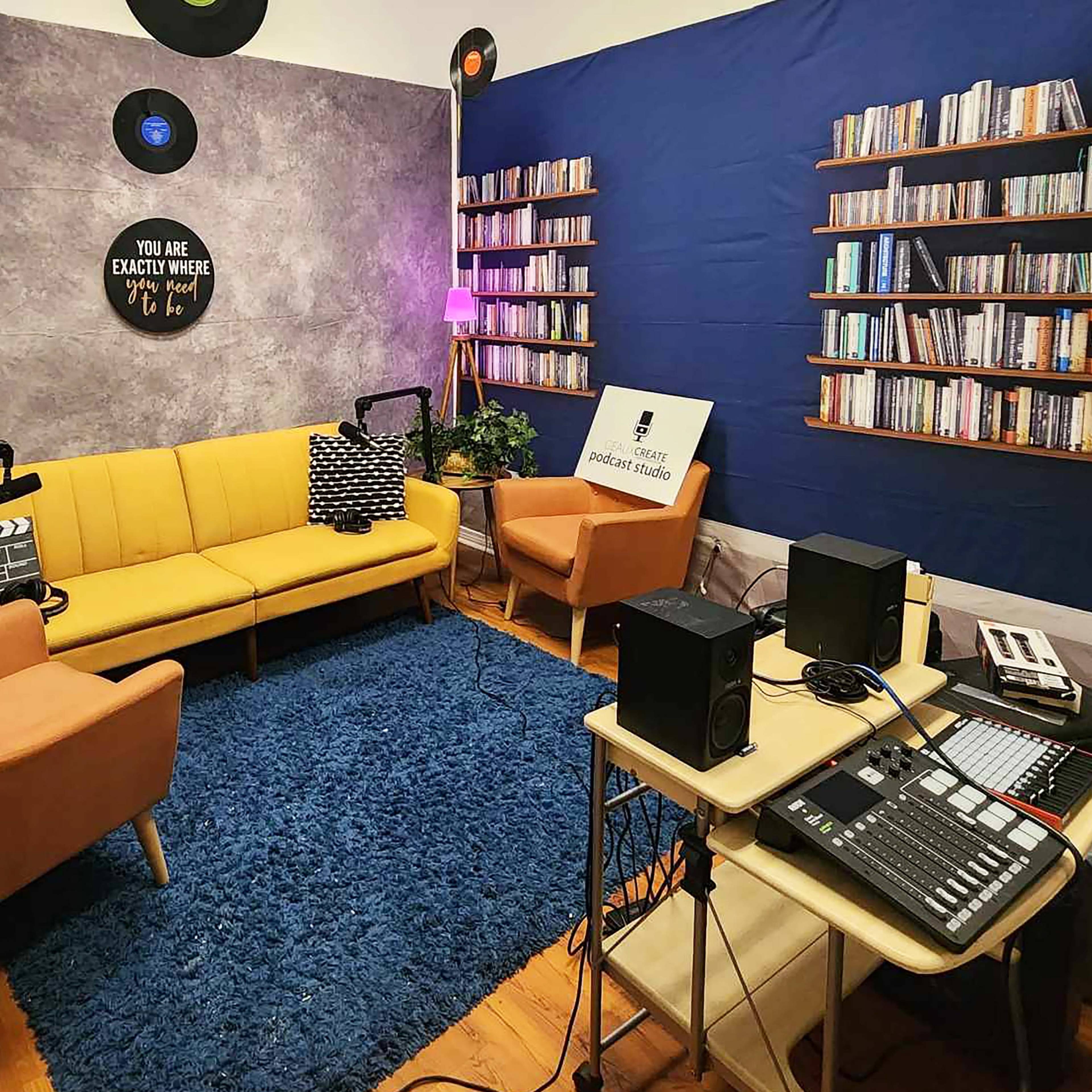 A music studio featuring a yellow sofa, two armchairs, a blue wall with bookshelves, and various audio equipment on a desk.