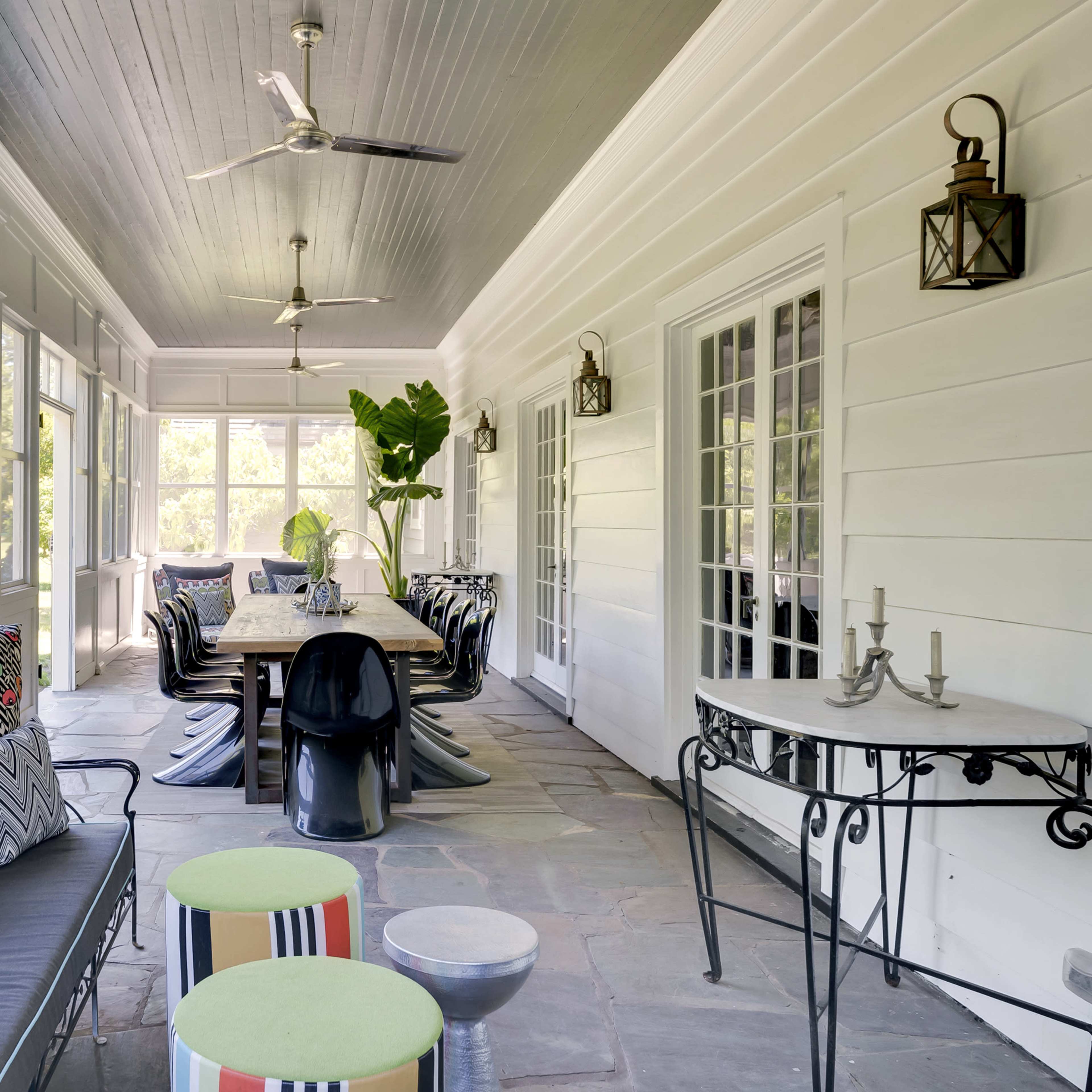 A covered porch with a long dining table surrounded by chairs, decorative cushions, and a stone floor.