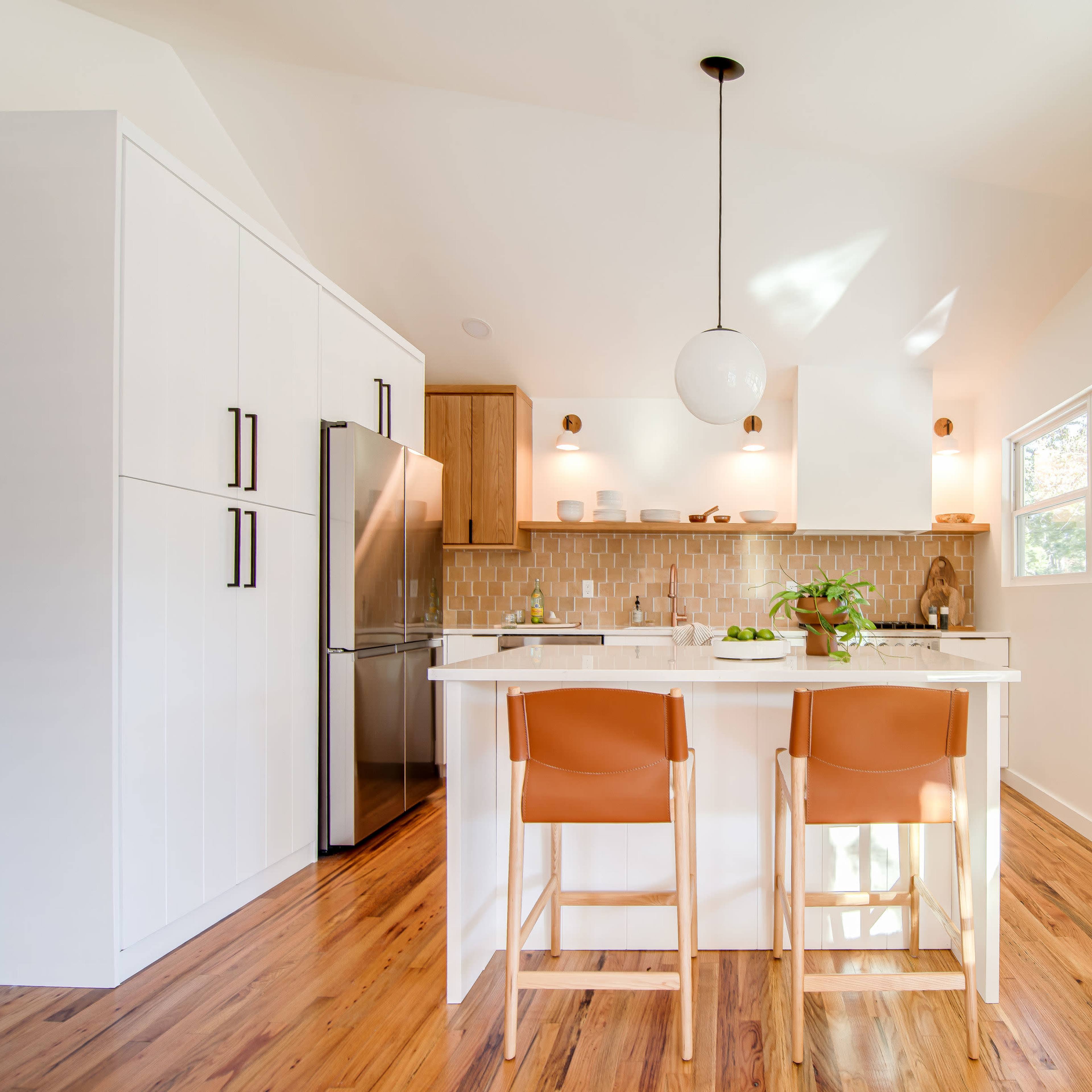 A modern kitchen features a white island with two tan chairs, a stainless steel refrigerator, and large windows that allow natural light to illuminate the space.