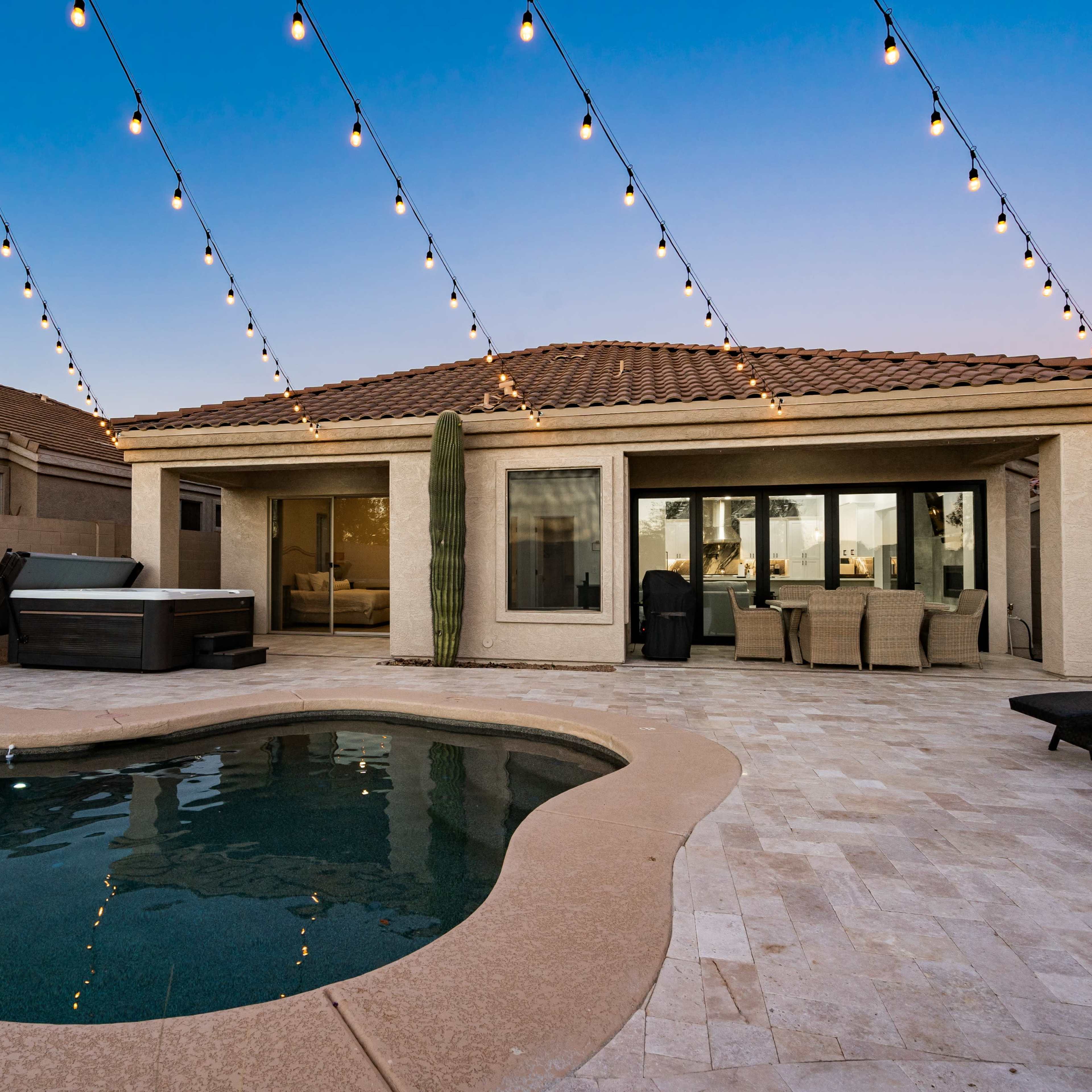 A backyard area with a pool, a hot tub, patio furniture, and string lights overhead, set against a twilight sky.