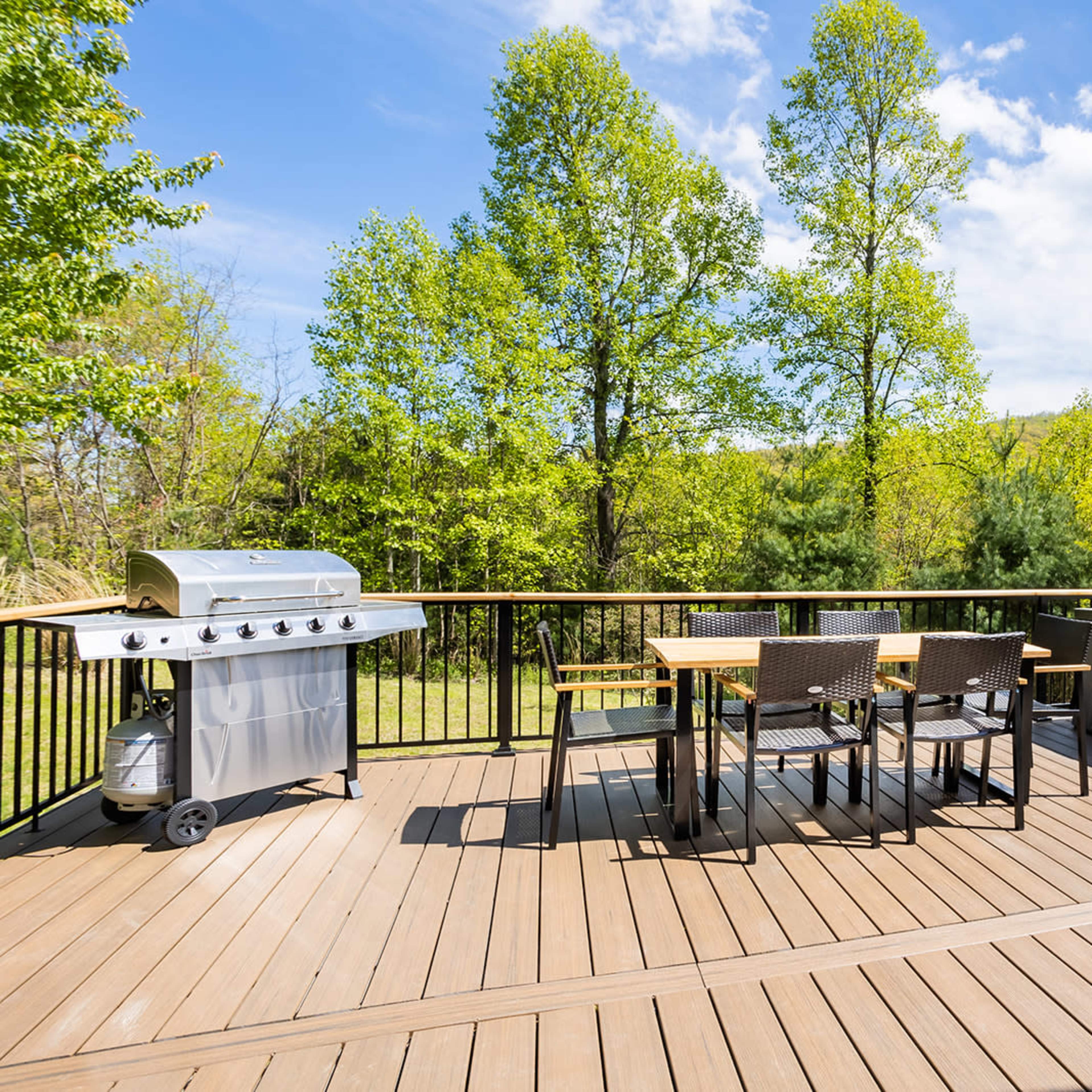 A wooden deck with a barbecue grill, a dining table with chairs, and a hot tub, surrounded by lush green trees under a partly cloudy sky.