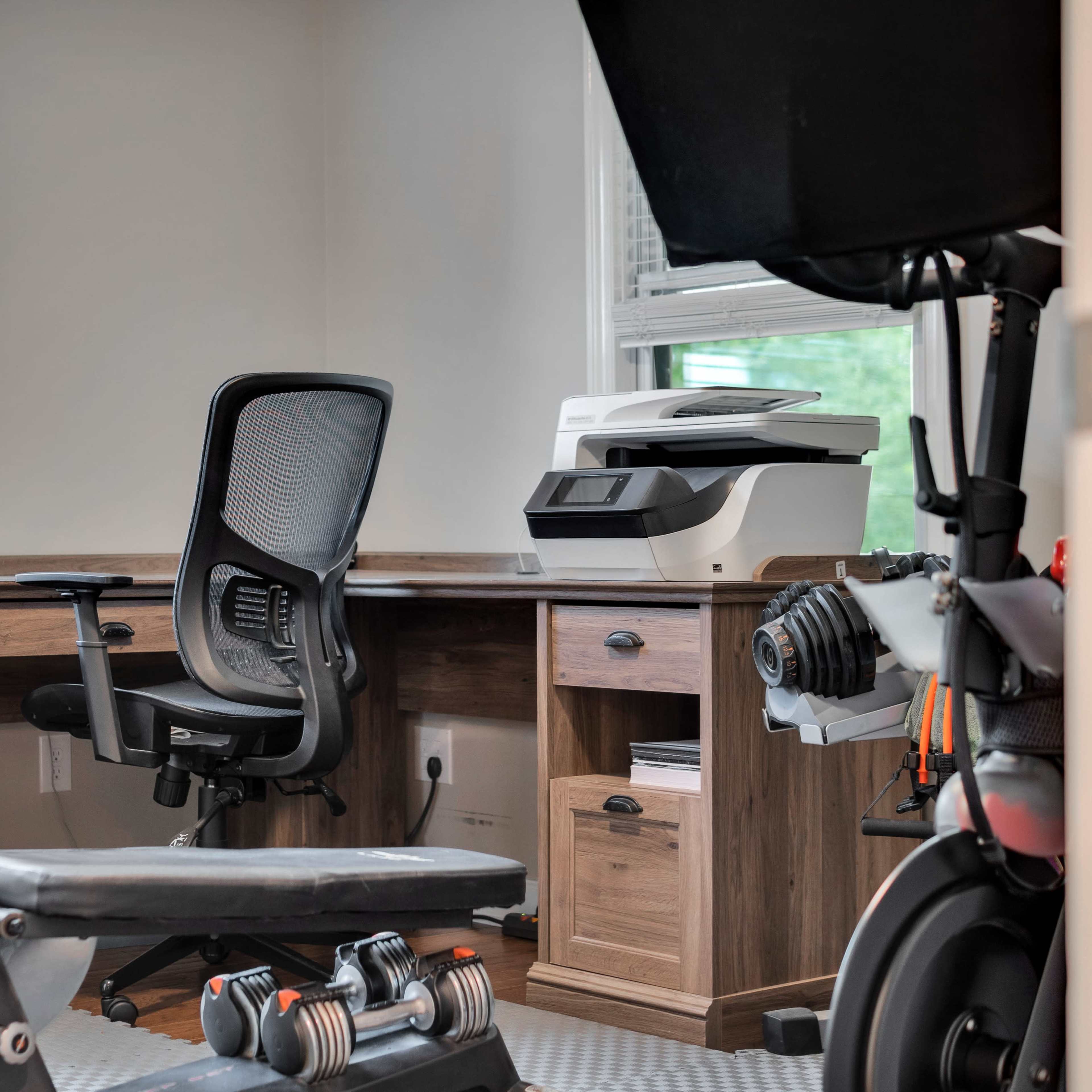 A home office space featuring a wooden desk with an ergonomic chair, a printer, and a workout bench with weights.