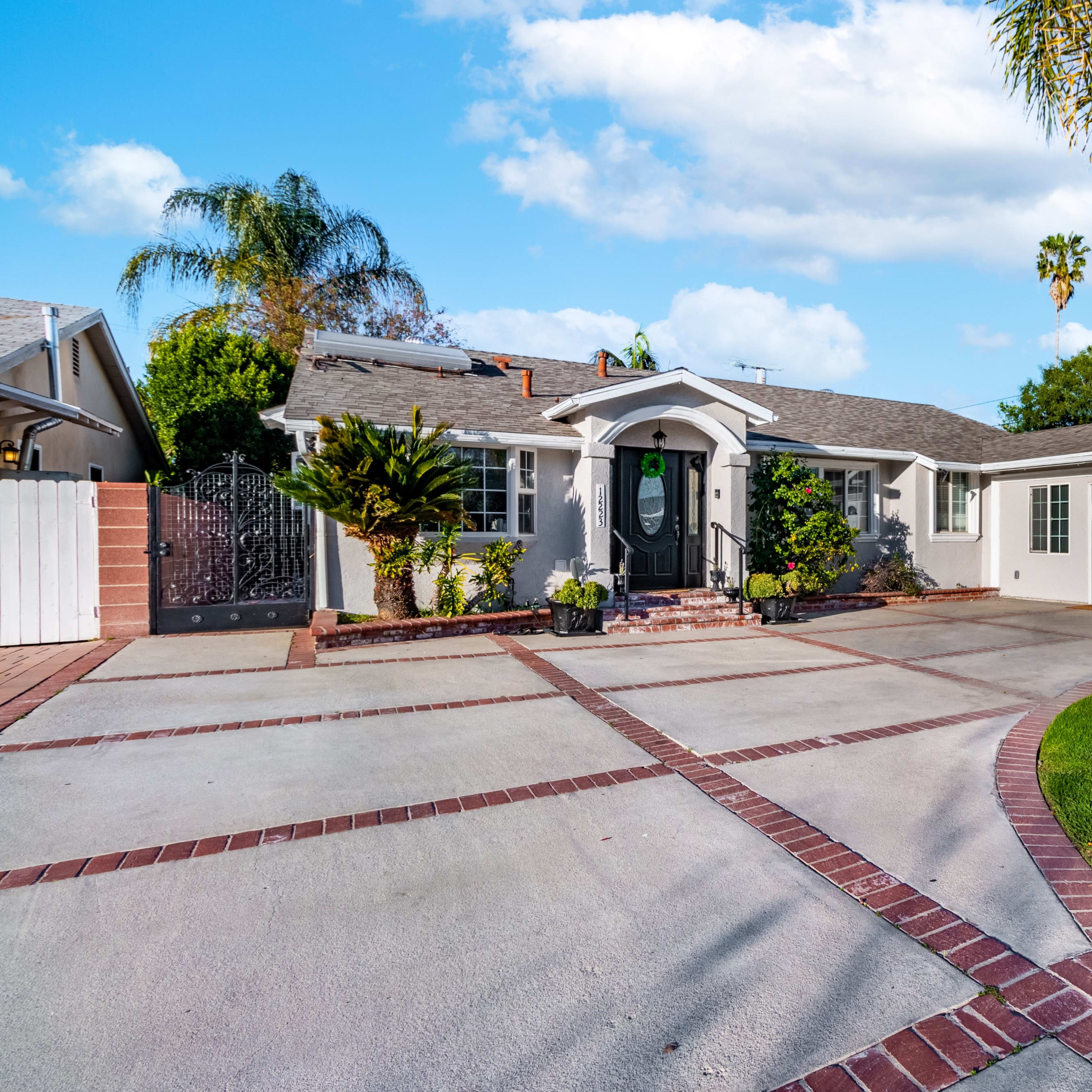 A single-story house with a manicured lawn and brick driveway features palm trees and a welcoming entrance.