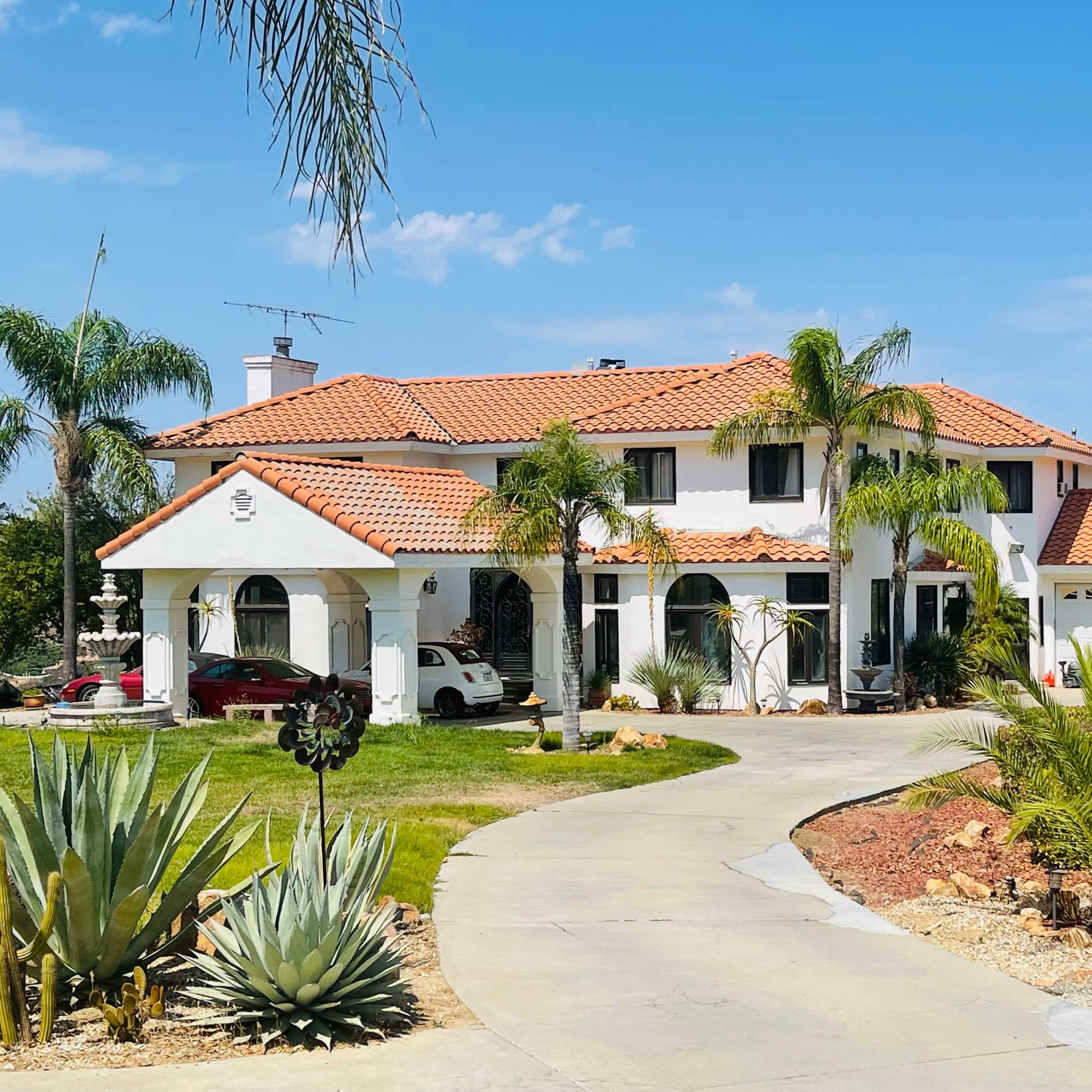 A large white house with a red tile roof, surrounded by palm trees and a circular driveway.