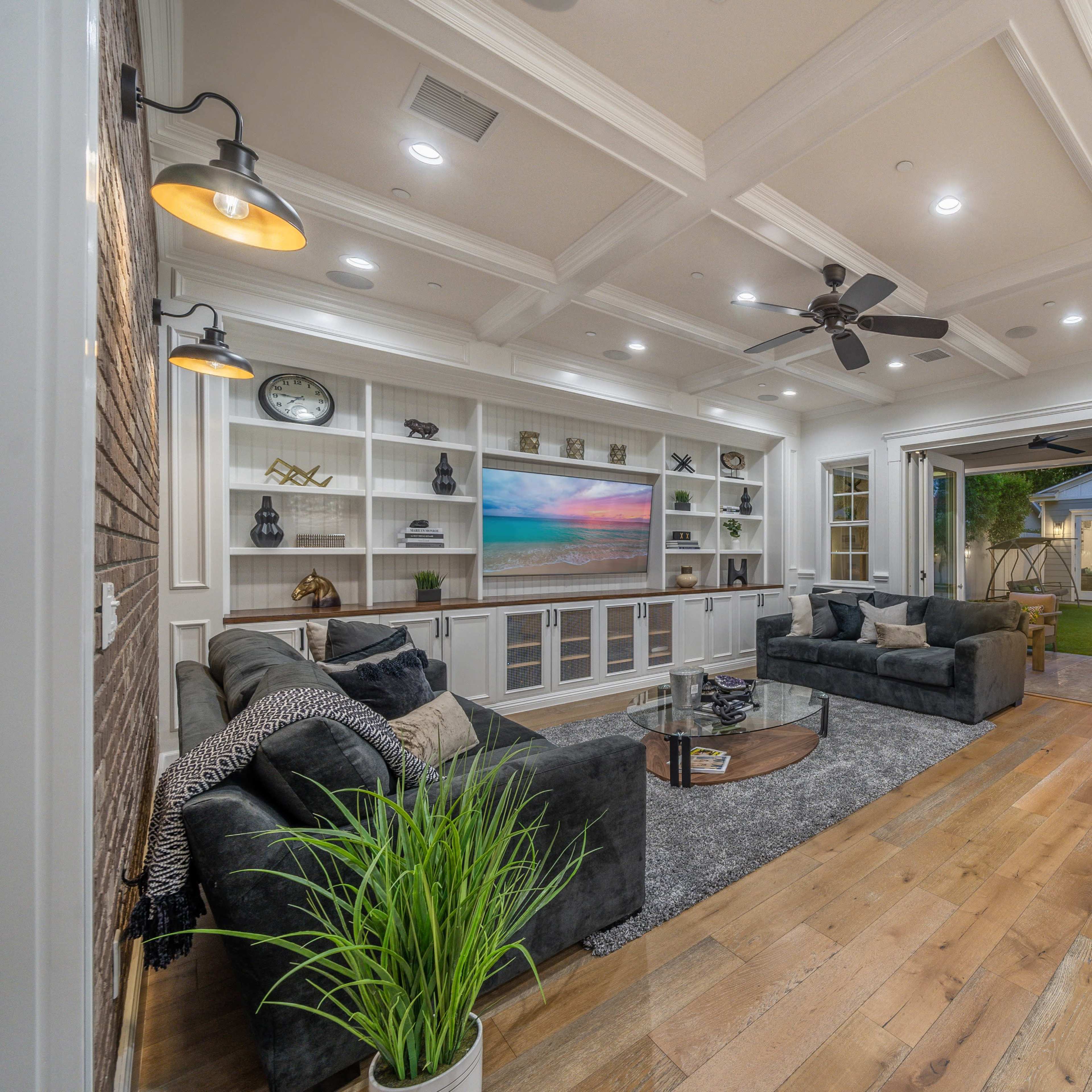 A modern living room with a coffered ceiling, a display cabinet, two sofas, and an open wall that leads to a backyard with a small shed.