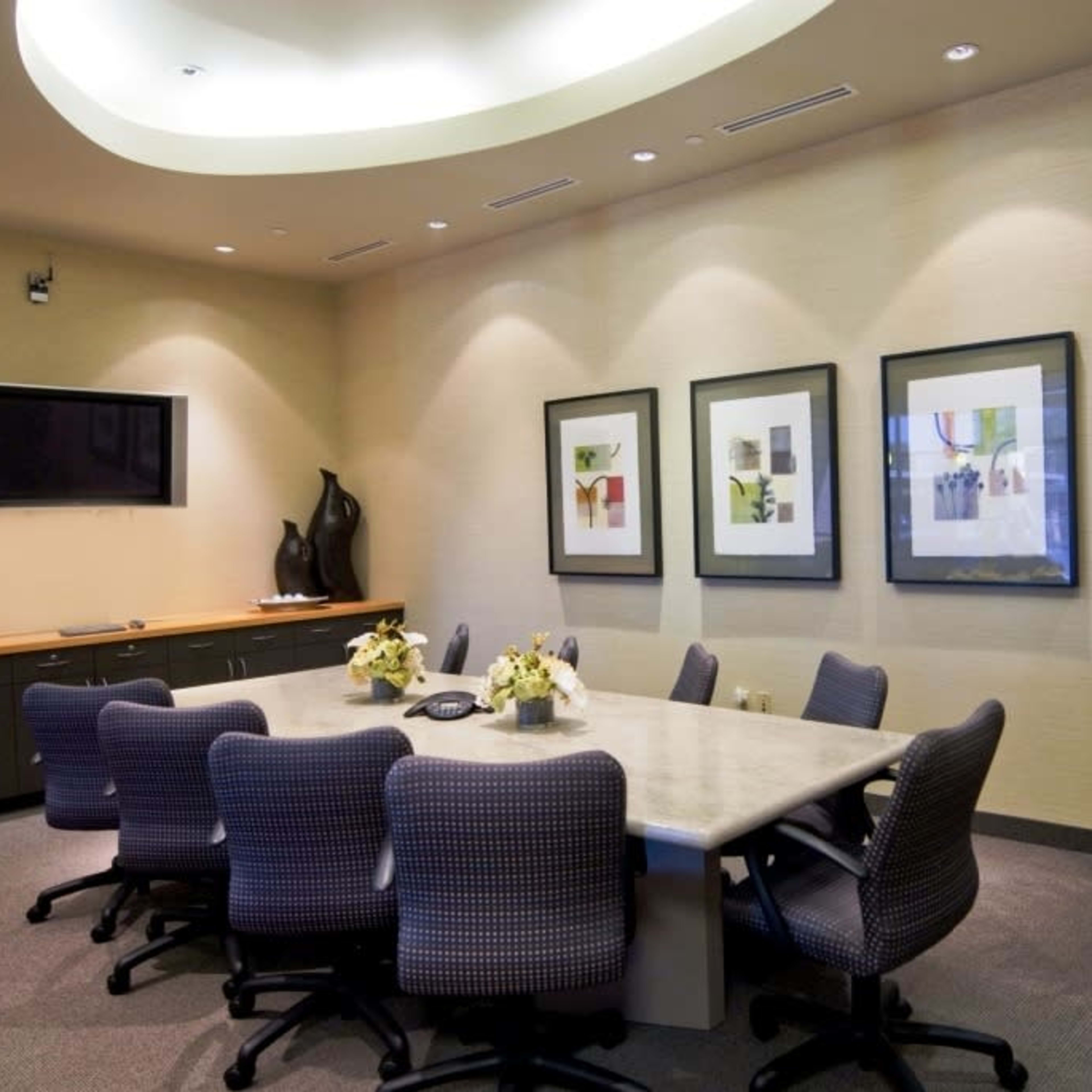 A conference room features a large table surrounded by several chairs and framed artwork on the walls, with a television mounted in the corner and a potted plant in the room.
