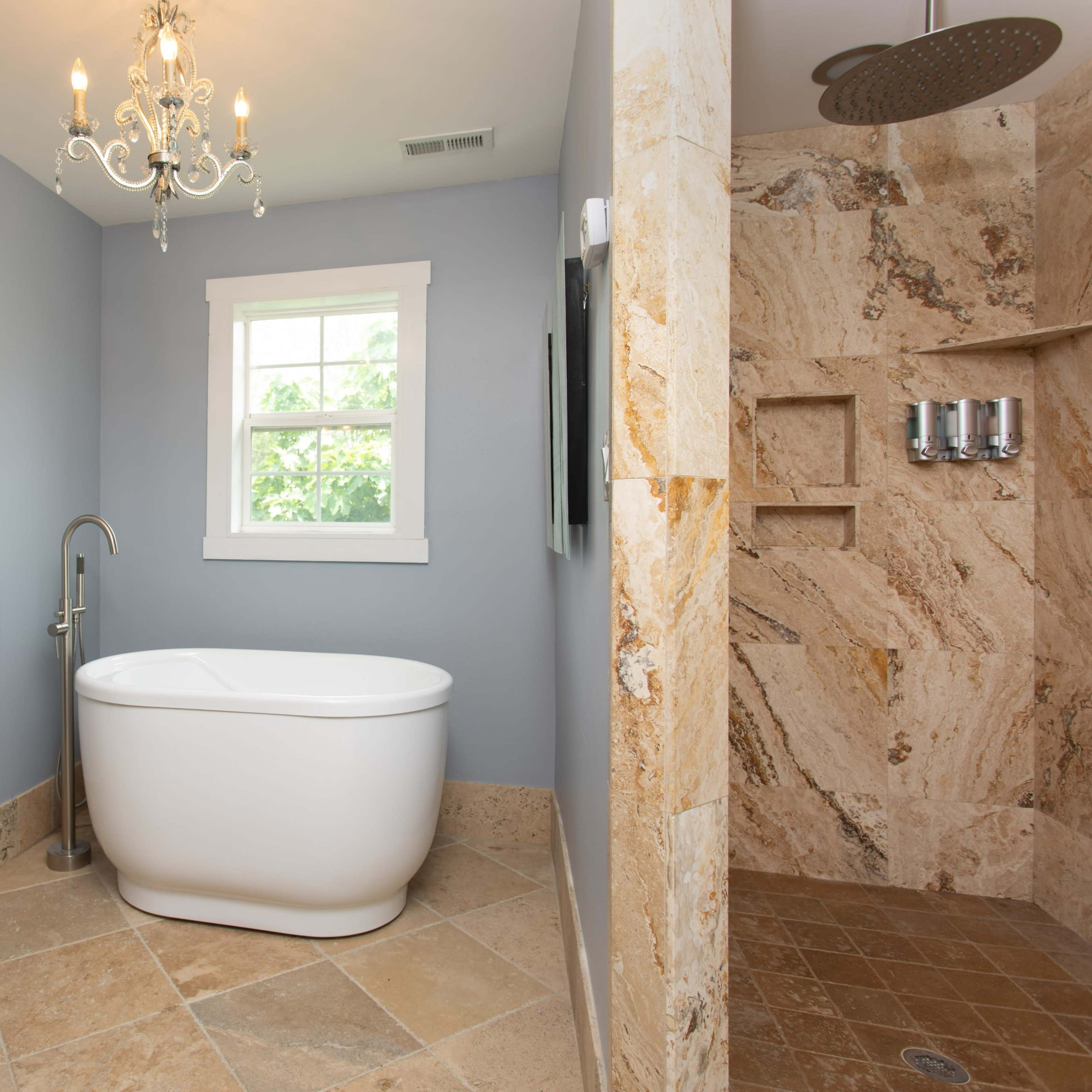 A bathroom featuring a freestanding bathtub, a stone-tiled shower, and a chandelier, with light blue walls and a window providing natural light.