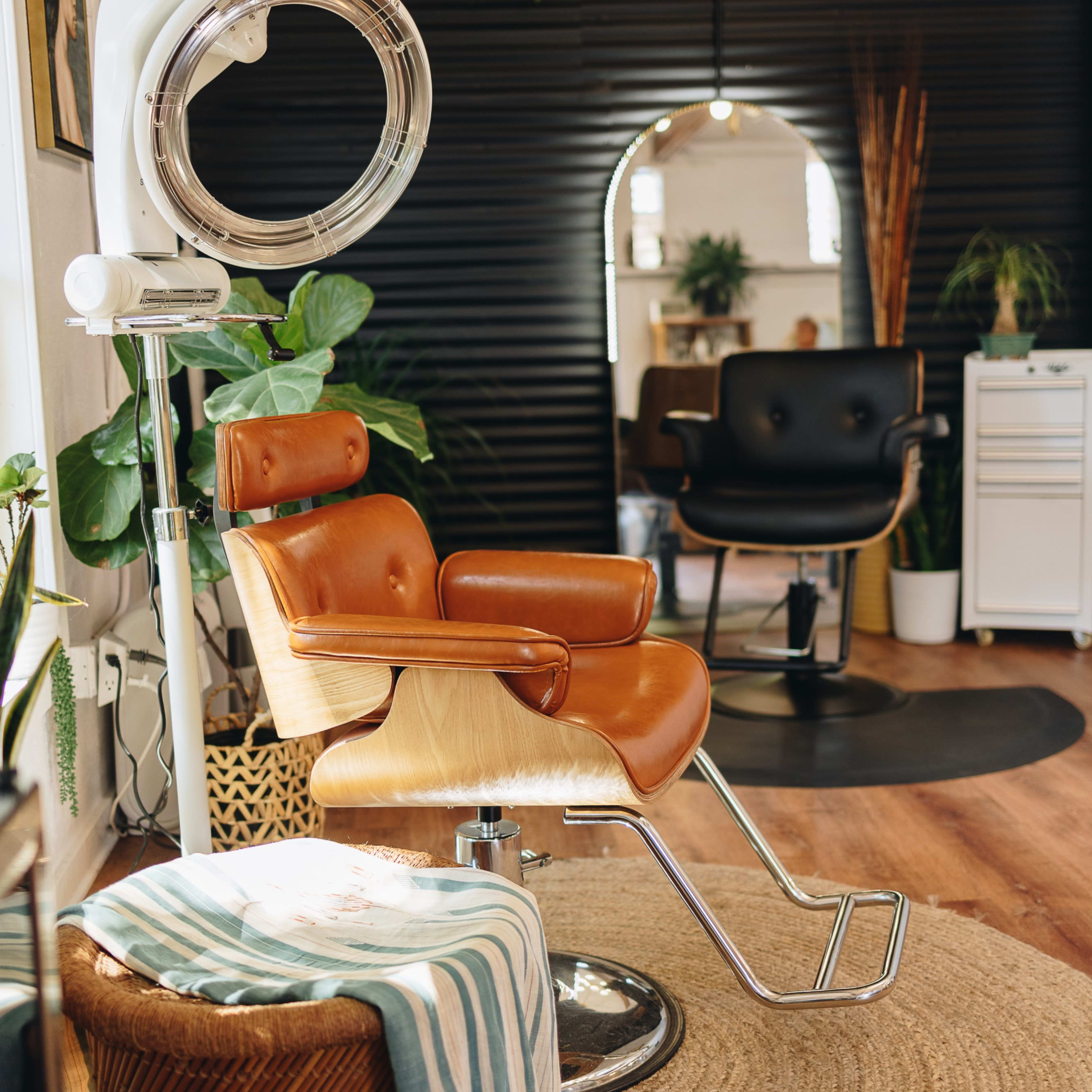 A modern hair salon interior featuring a brown leather styling chair, a round mirror, and lush green plants.