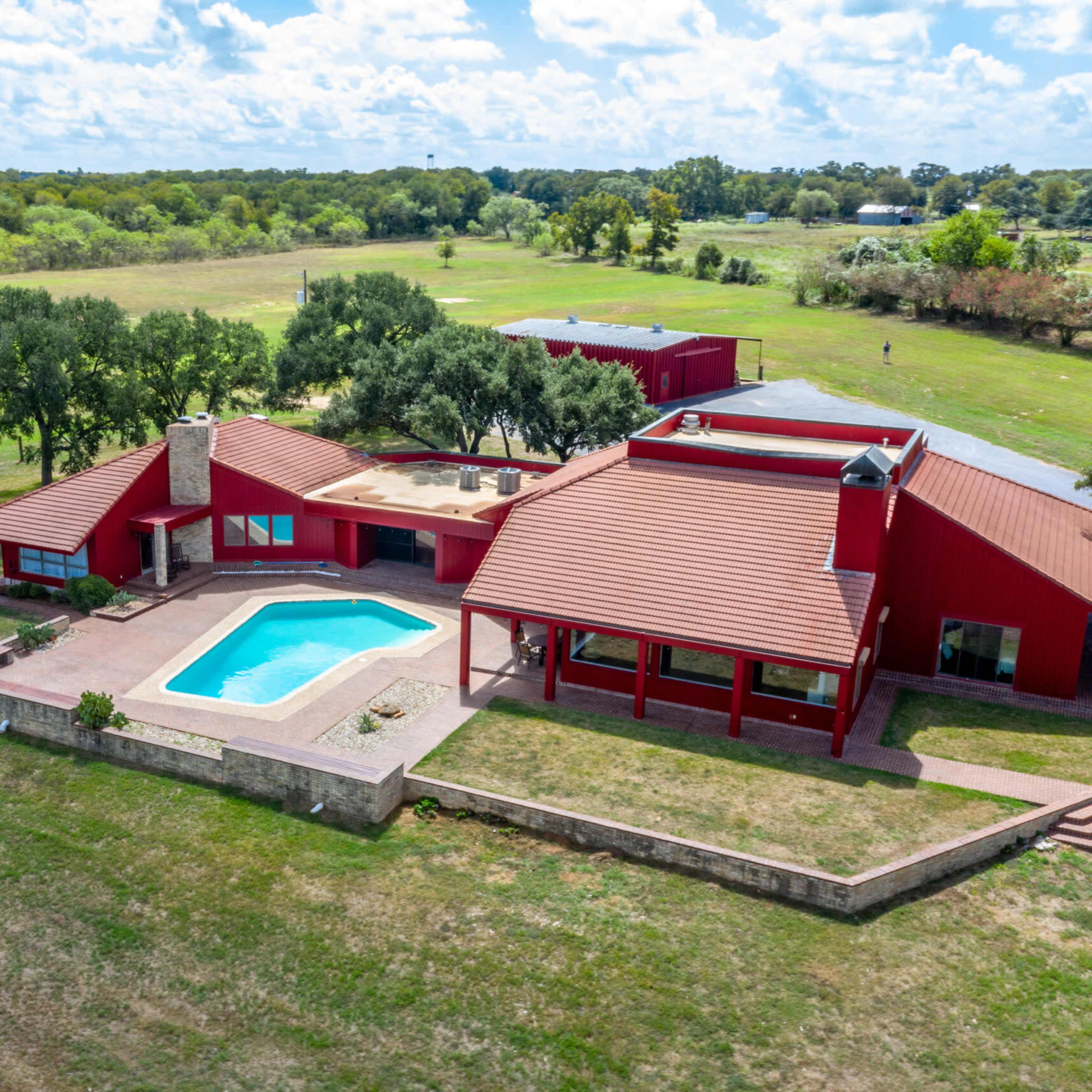 A red-stucco house with a swimming pool is surrounded by green fields and trees under a cloudy sky.