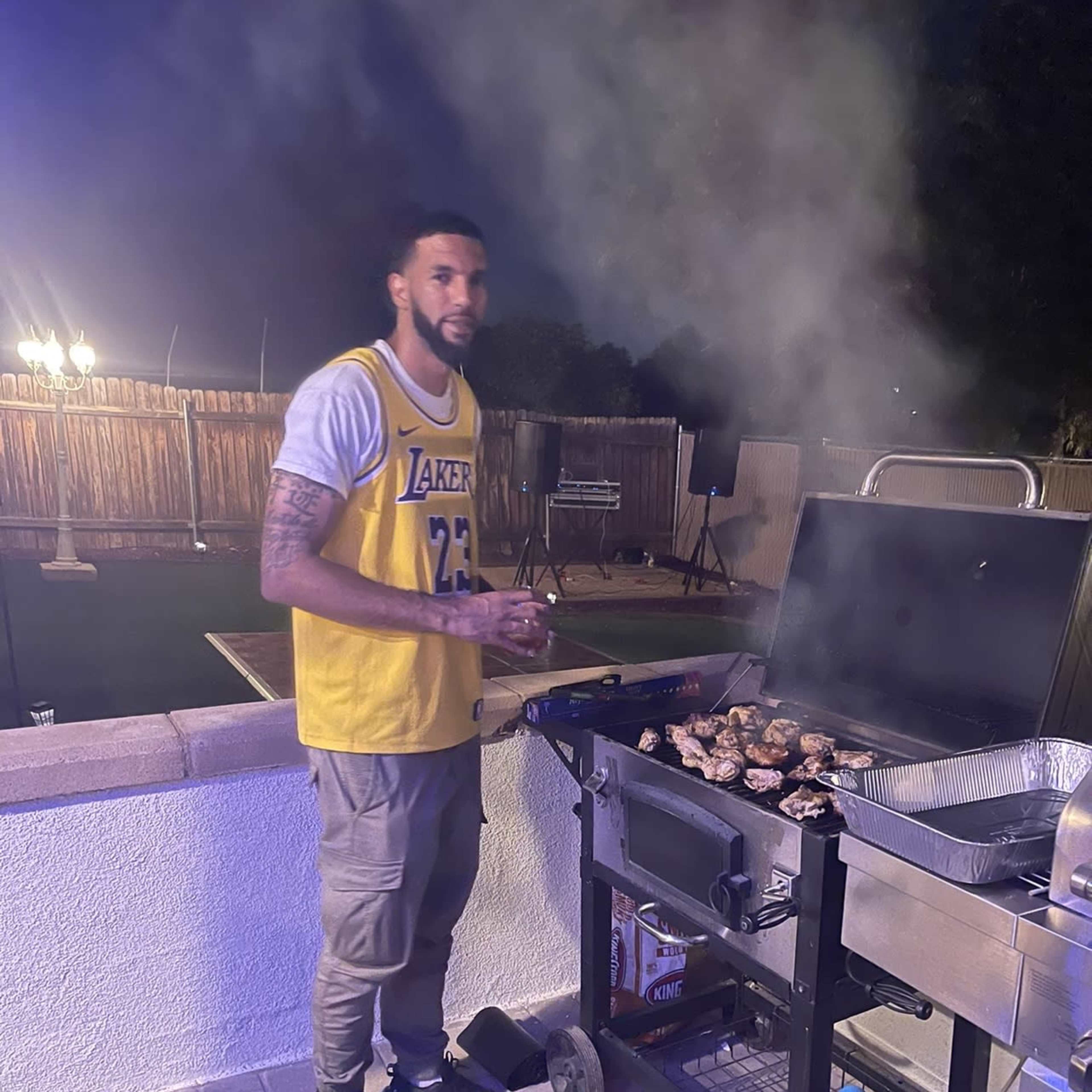 A man stands next to a grill outdoors at night, cooking chicken while surrounded by smoke.