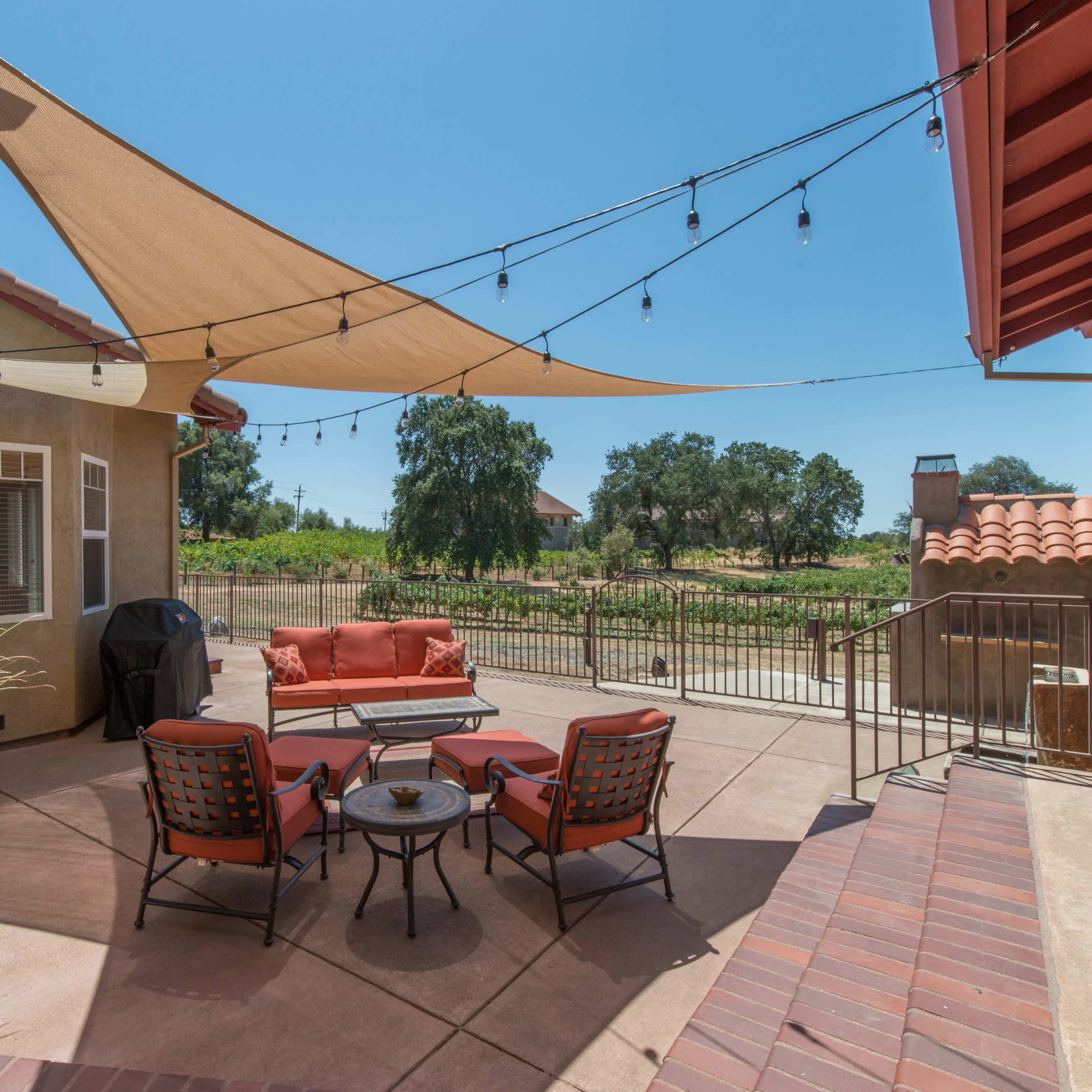 A patio area with a seating arrangement, shaded by a sail, overlooks a vineyard.