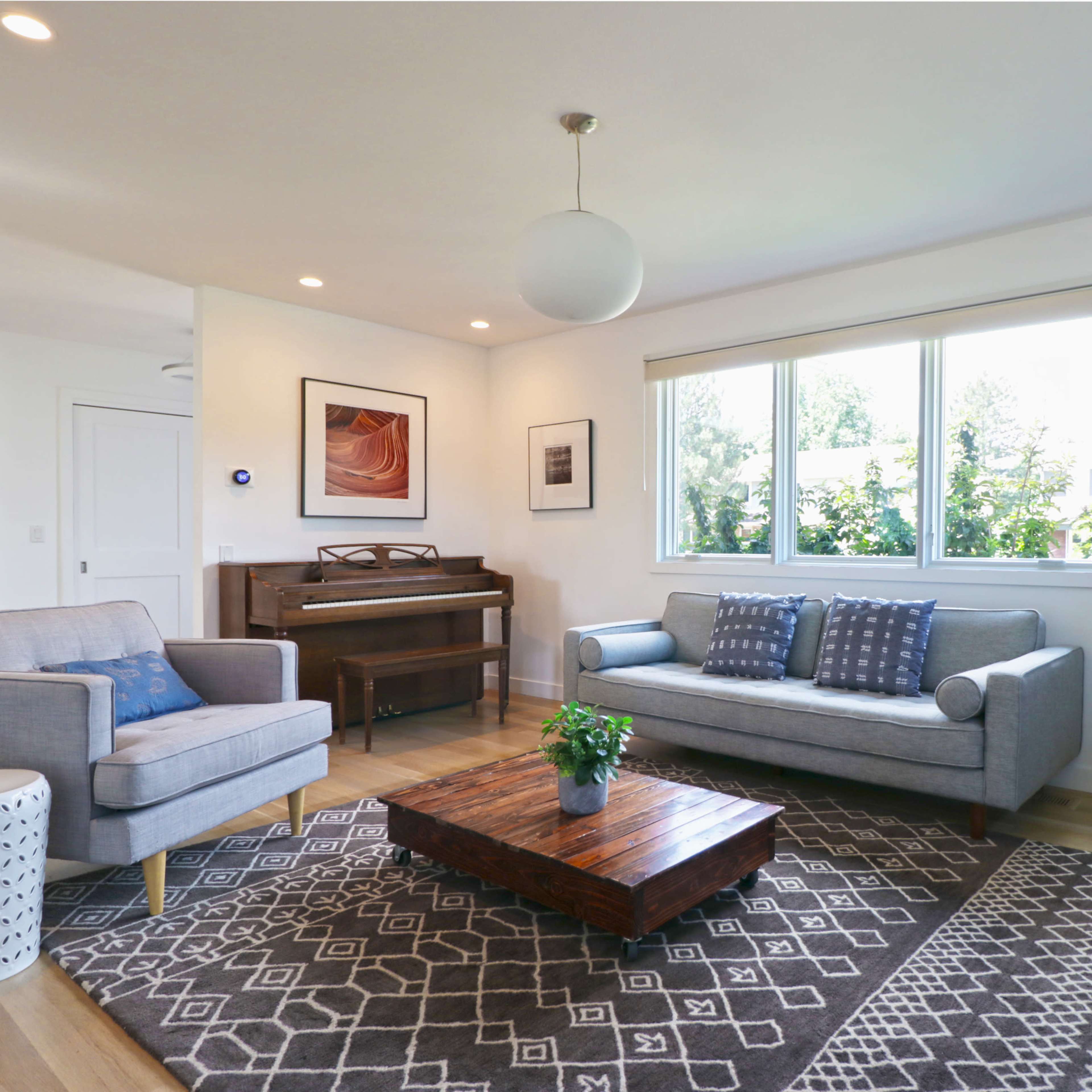 A bright and modern living room features a piano, two gray sofas, a wooden coffee table, and large windows offering a view of greenery.